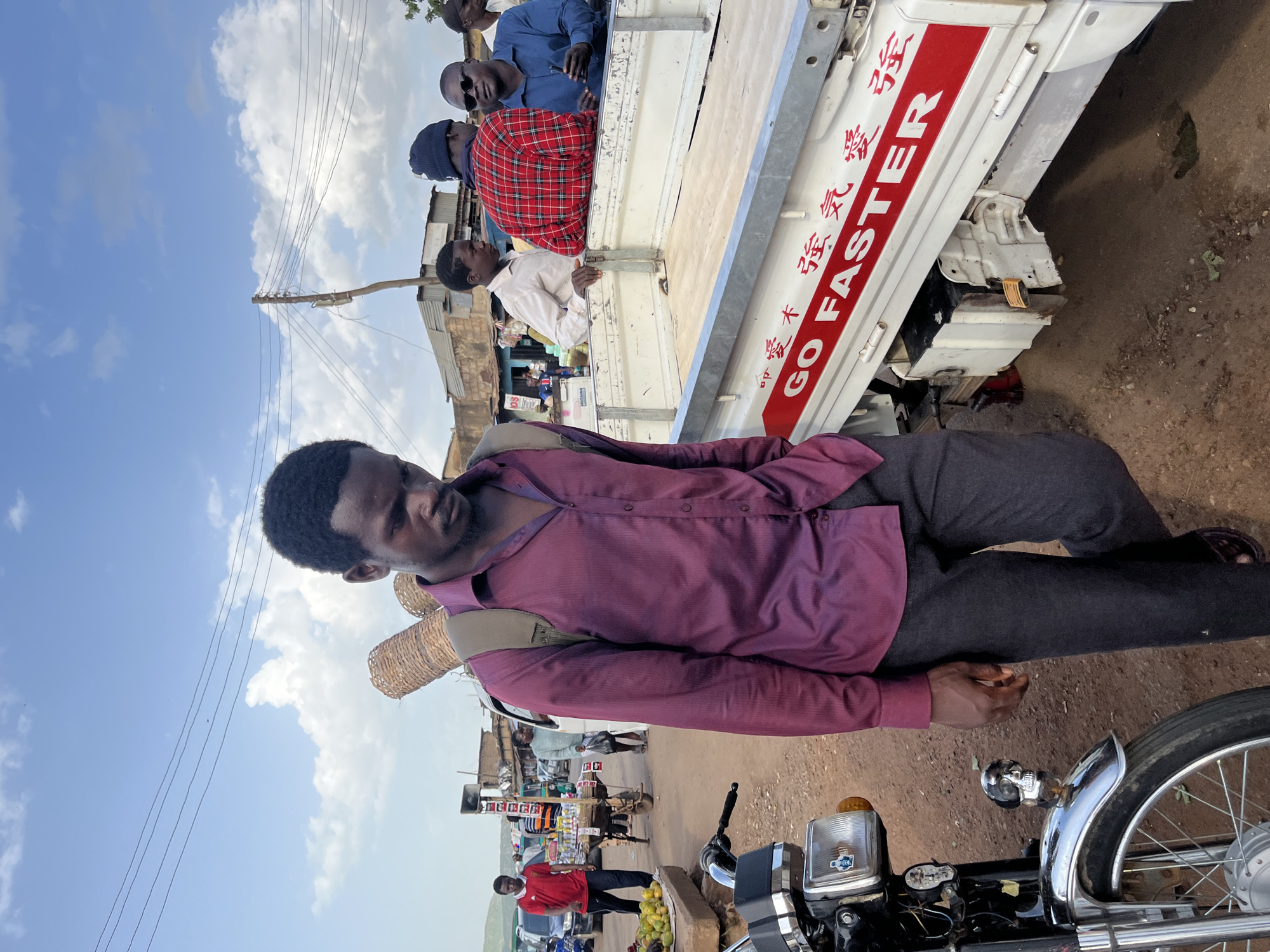 The media file is a portrait of a man in Rafin Par, Nigeria. The man is wearing a purple button-up shirt and dark pants, with a woven basket slung over his shoulder. He is standing in front of a white truck with red lettering that reads "GO FASTER" in English and Chinese characters. The truck appears to be carrying several passengers in its bed.
The scene suggests a market or a busy roadside area. In the background, there are buildings, a power pole with wires, and other vehicles, including a motorcycle in the foreground. The weather appears to be sunny with some clouds in the sky. The time of day seems to be daytime, likely late morning or early afternoon. The man's expression is neutral, and the overall mood of the image is that of everyday life in a bustling environment. The presence of the truck with people indicates transportation and community activity. The man's attire and the basket suggest he might be a vendor or a traveler carrying goods. The setting, with its unpaved ground and simple structures, points to a rural or semi-urban area.