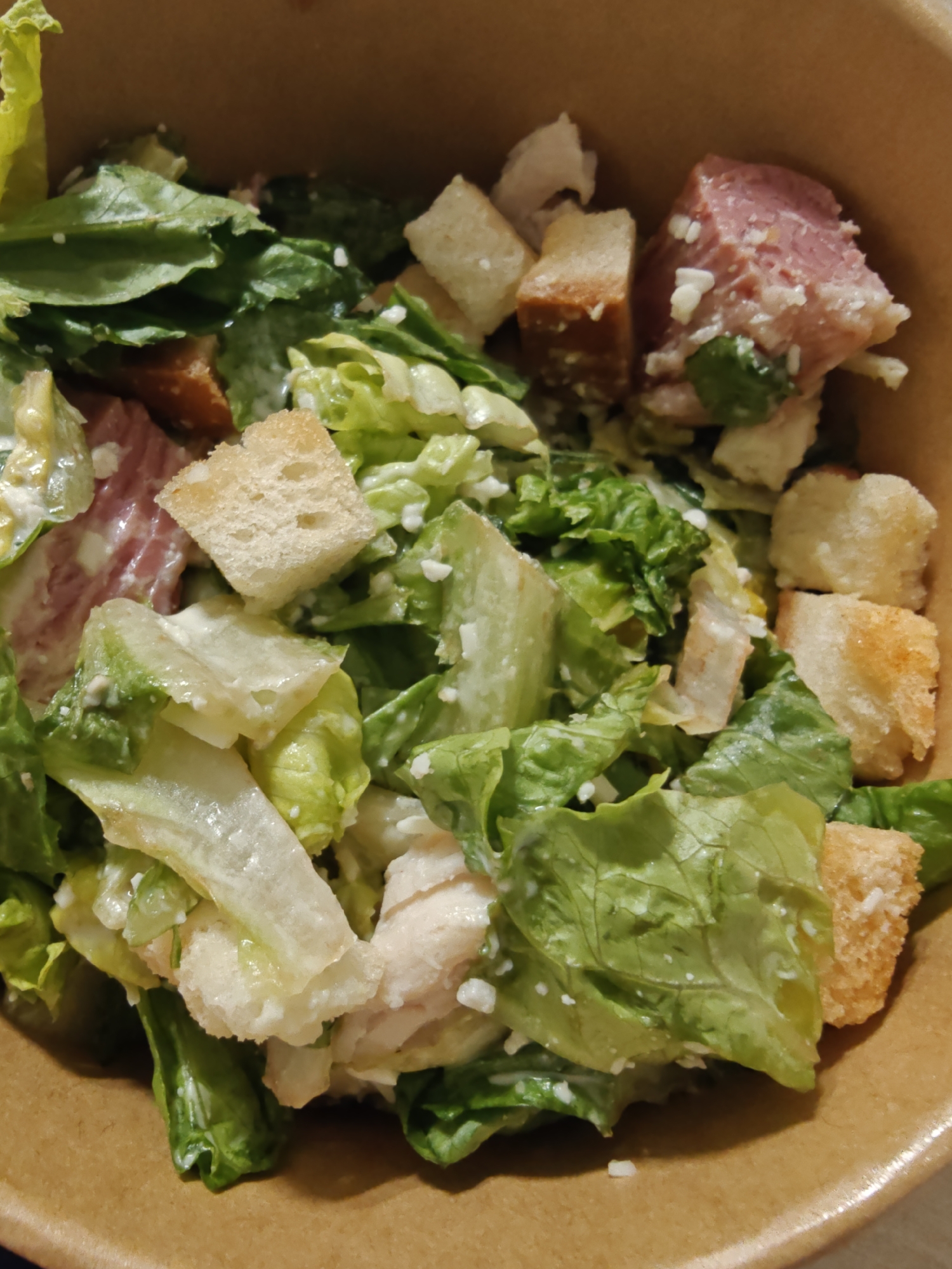 The image shows a close-up, overhead view of a Caesar salad in a cardboard bowl. The salad contains chopped romaine lettuce, croutons, and chunks of what appears to be ham and cooked chicken, all coated in a creamy dressing. There are also flecks of white cheese visible throughout the salad. The salad is presented in a light brown, textured cardboard container, suggesting it might be takeout or a casual dining setting. The lighting is dim, and the focus is on the food, with the background slightly blurred. The image does not contain any information about a specific location in Dubai or the United Arab Emirates.