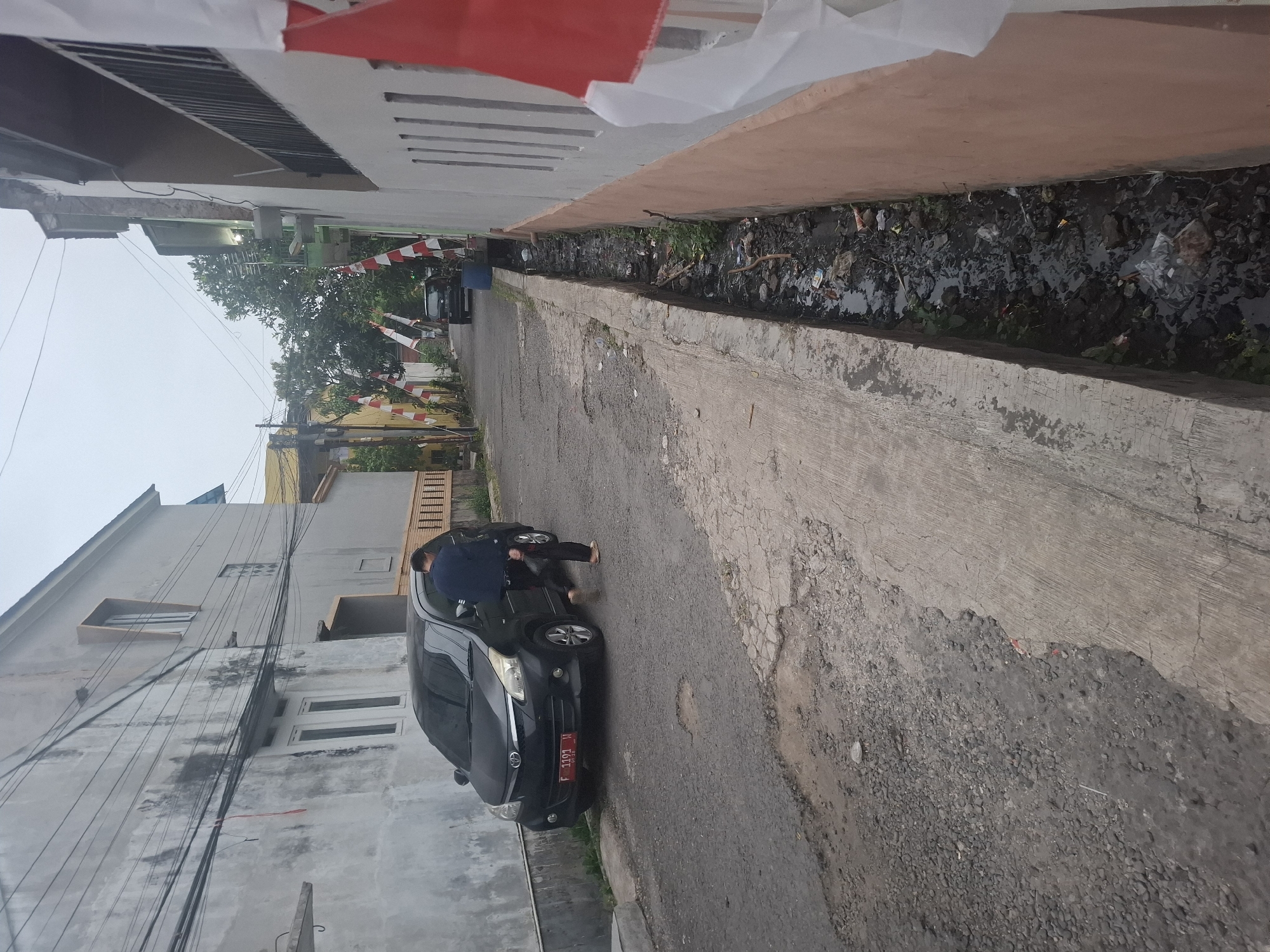 In the city of Karangtengah, Indonesia, an overcast day presents a narrow, paved street lined with buildings and a drainage ditch. A dark-colored car, a Toyota Avanza with license plate F 1193 W, is parked on the left side of the road. A person, wearing a dark blue jacket and dark pants, stands next to the driver's side door of the car, facing away from the camera, with one foot on the ground and the other appearing to be in the process of stepping out or in. Red and white Indonesian flags are intermittently displayed along the street. The drainage ditch on the right side of the street is filled with murky water, debris, and what appears to be trash. The road surface is uneven and shows signs of wear. Buildings on both sides of the street are of varying heights and architectural styles, with one on the left exhibiting external electrical wiring. The overall scene suggests a residential area.