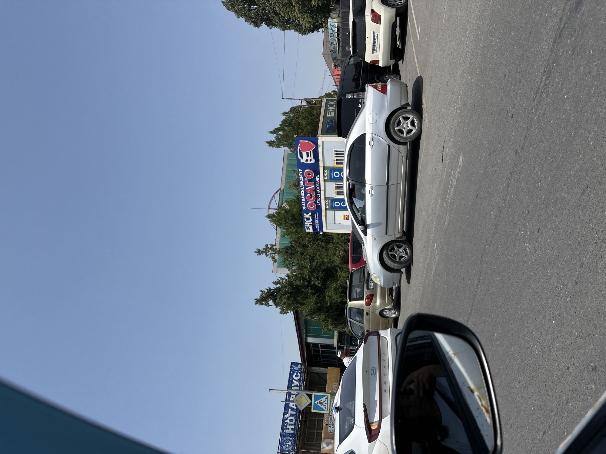This is a photograph taken from a car, showing a street scene with several vehicles and buildings. The location is identified as the city of Manas, Kyrgyzstan. In the foreground, a white car is visible, with its side mirror reflecting a person taking the picture. Behind it, a silver sedan is in motion, followed by a black van and a white SUV. Further back, a small gold car and a red vehicle are parked. Buildings with signage are visible in the background, including "NOTARIUS" and "OSAGO" with a shield logo. The sky is clear and blue, indicating a sunny day. The road appears to be asphalt.
