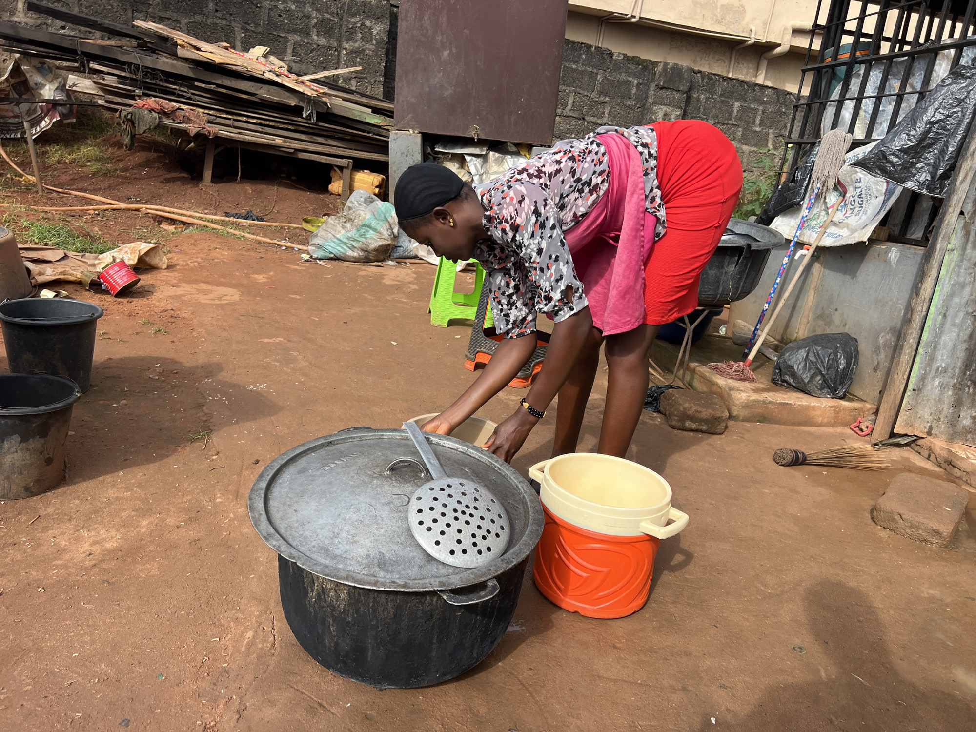 In Nkpor, Nigeria, a young woman is actively engaged in food preparation outdoors. She is bending over a large, black cooking pot with a metal lid, using a slotted metal spoon to handle food. She wears a patterned black, white, and pink blouse, a bright red skirt, and a black head covering. Beside the large pot, an orange and cream-colored container sits on the ground. The environment appears to be a dusty, outdoor area with a rough, reddish-brown dirt ground. Scattered around are various objects, including black buckets, sacks, wooden planks, a broom, and a mop. The background shows a brick wall and some corrugated metal structures, suggesting a residential or commercial outdoor workspace.