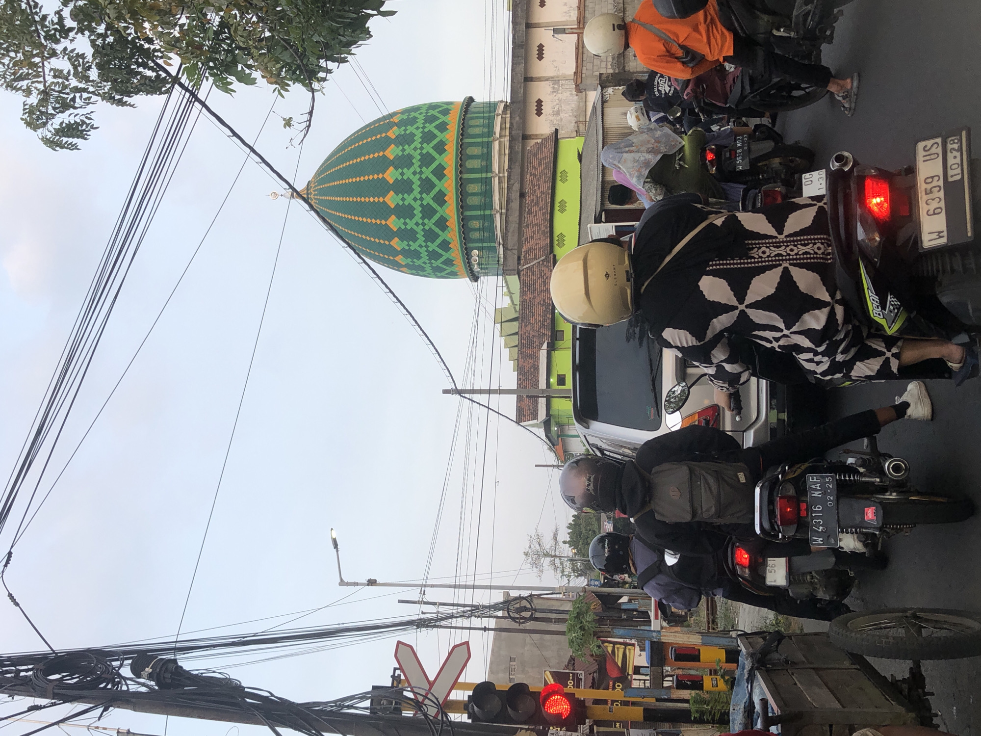 The image depicts a street scene in Sumorame, Indonesia, characterized by a traffic jam and several motorcycles. In the background, a green mosque with an orange-trimmed dome stands prominently. Power lines are visible crisscrossing the sky, and a traffic light showing red is present on the left side of the frame. Several individuals wearing helmets are riding motorcycles, some with passengers. One motorcycle has a license plate reading "W 4316 NAF" and "02.25". Another motorcycle has a license plate reading "W 6359 US" and "10.28". A wooden cart with one visible wheel is parked on the left. The environment appears to be an urban or suburban setting.