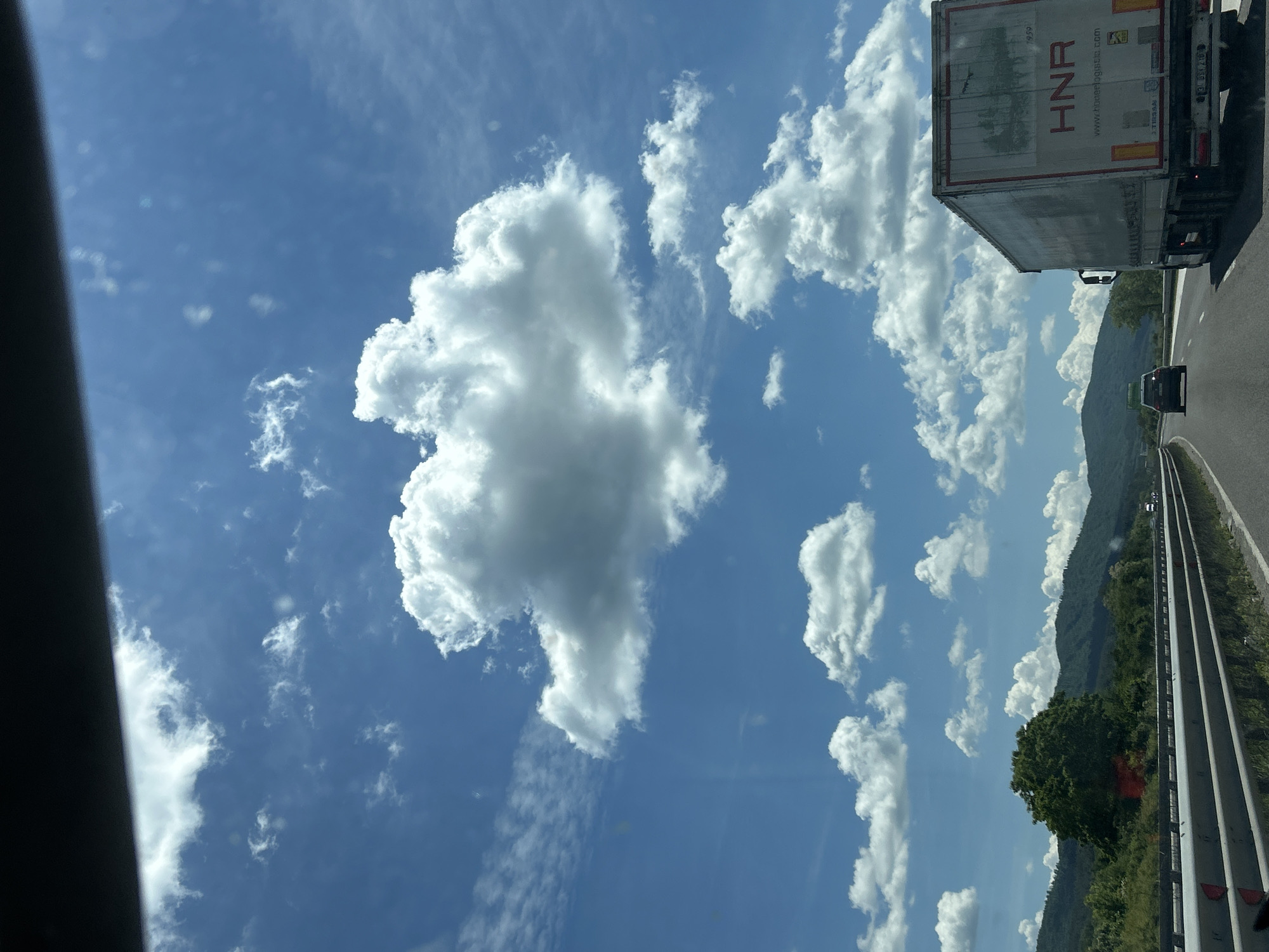 The image depicts a view from a vehicle traveling on a highway in Verinsko, Bulgaria, under a bright, cloudy sky. A large, white truck with the company name "HNR" and website "www.hnrlogistics.com" is prominent in the foreground, to the right. A dark-colored car is visible ahead on the highway, to the left of the truck. The highway is bordered by guardrails and a grassy verge. In the background, lush green hills covered in trees rise under a sky filled with fluffy white cumulus clouds and wispy cirrus clouds. The overall scene suggests a sunny day on a road trip through a scenic, rural area.