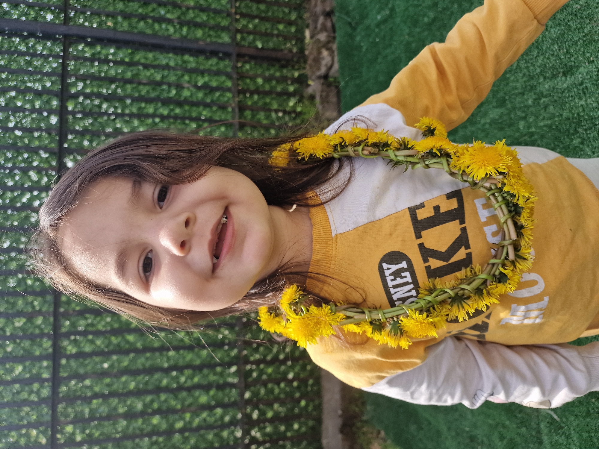 A young child with long brown hair is smiling at the camera. The child is wearing a yellow and white long-sleeved shirt with a graphic print. A wreath of yellow dandelions is worn around their neck. The background features a dark metal fence with greenery behind it and an area of green artificial turf or grass visible to the side. The location appears to be outdoors, but the city is Unknown, Unknown.