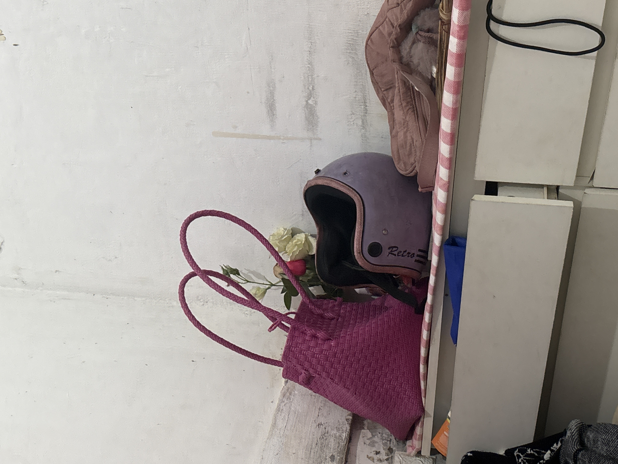 A purple helmet and a pink woven bag sit on a shelf in a room. A pink checkered cloth covers the shelf. A light pink puffy bag is next to the helmet. The background is a white wall. The shelf is above two white drawers.