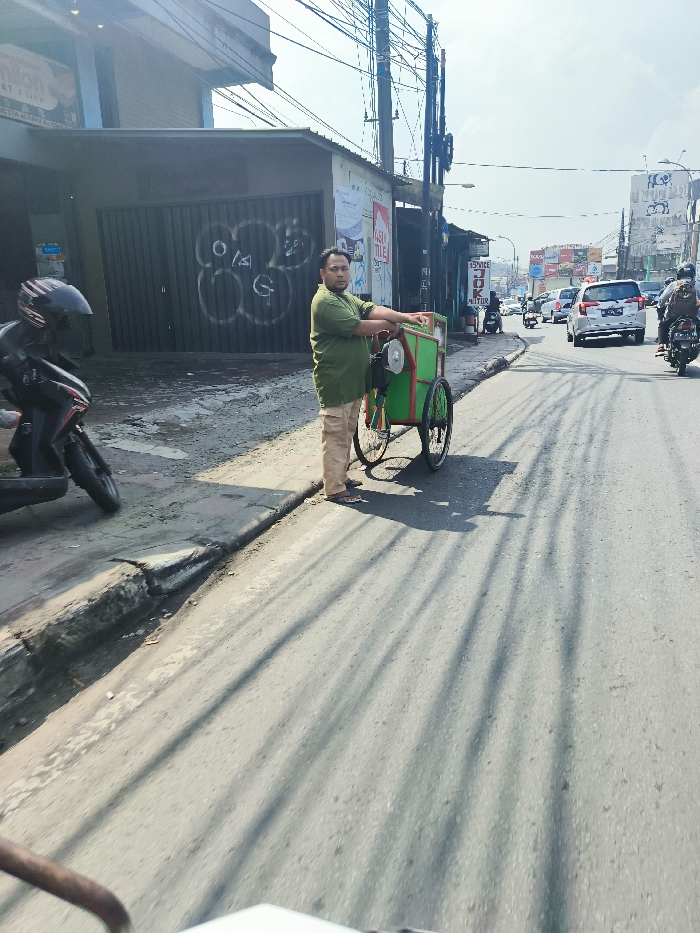 A man in a green shirt and tan pants stands next to a small, green, and yellow food cart in a street in Cileunyi, Indonesia. The cart is being loaded with food items. The man is facing away from the camera, but his face is visible. The cart has two wheels and is in the middle of the street. There are several other vehicles parked in the background, and one car is driving toward the camera. The road is paved with asphalt, and the sidewalk is lined with a row of small bushes. The man is looking down at the cart, and it is not possible to see his face. The sky is partly cloudy and sunny. The street is lined with buildings, and the image is taken from the perspective of a vehicle driving down the street. There are several electrical poles and wires in the background. The road is empty except for the man and the cart. The man is alone. He is about to push the cart. The road is in a residential area. The image was taken during the day. The light is good. The camera is pointing toward the front of the man and the cart. The street is relatively narrow, and there is a sidewalk on the right side of the street. The sidewalk is made of concrete and is covered with a thin layer of dirt. The buildings on the left side of the street are old and worn. The buildings on the right side of the street are modern and well-maintained. The area is a mixture of residential and commercial property.