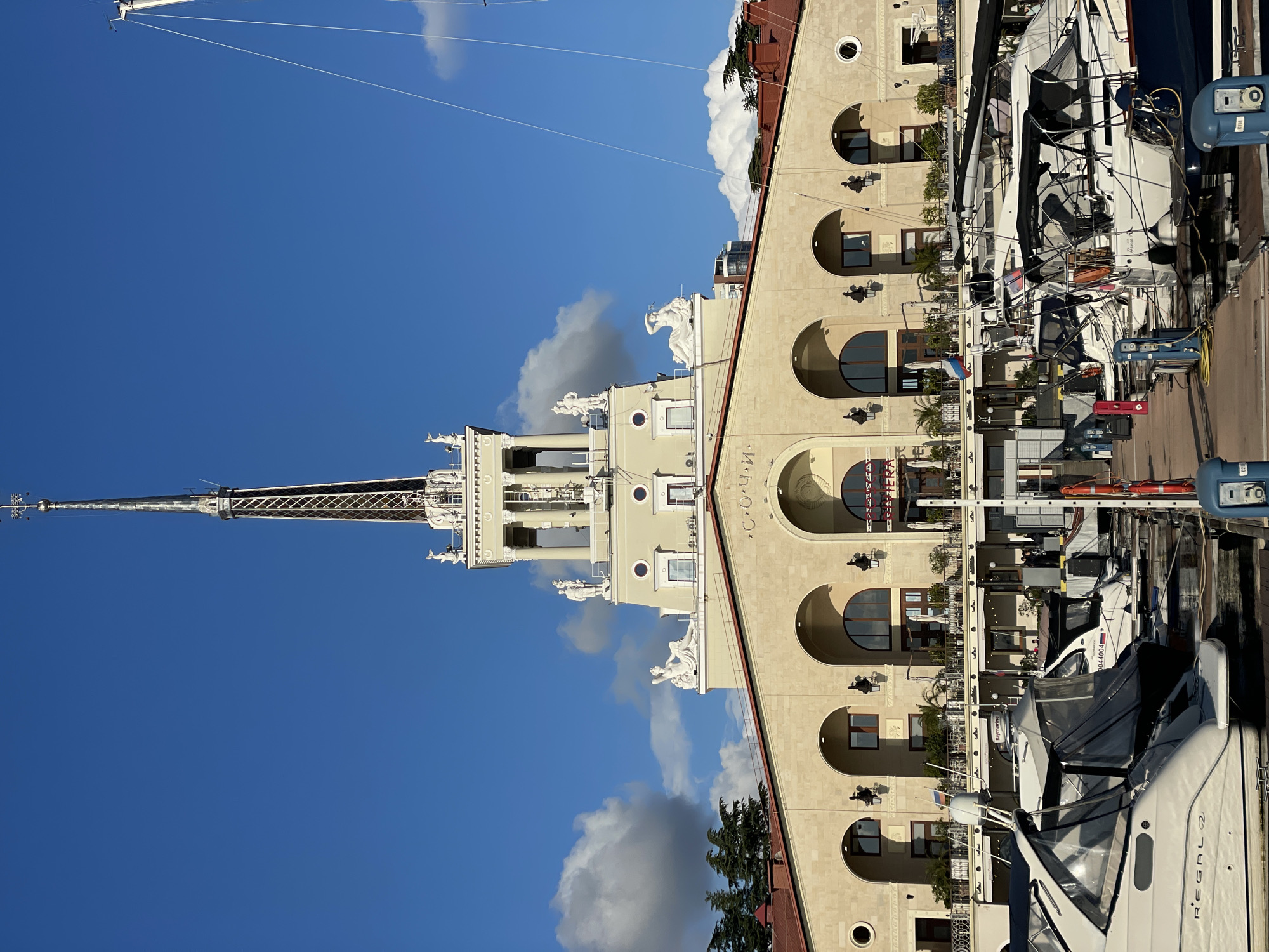 A vertical shot shows a historical building with a spire, partially obscured by moored yachts along a waterfront. The building is beige, with arched windows and decorative statues near its top. The spire is ornate, and visible against a blue sky with scattered clouds. The yachts obscure the base of the building. Located in Sochi, Russia, the scene suggests a marina or port area. The shot is taken from a ground-level perspective.