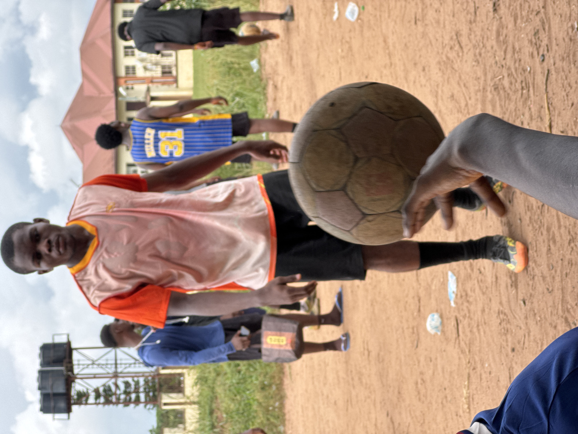 A vertically oriented, candid shot captures a group of young Black men in an outdoor, unpaved area, likely in Ihiagwa, Nigeria, during the daytime under an overcast sky.
In the foreground, a young man wearing a light orange/peach sleeveless shirt with orange trim and dark shorts looks directly at the camera with a neutral expression. His arms are bent, suggesting a paused motion or relaxed stance. To the right, a weathered, light brown soccer ball is held by an unseen person's hand, positioned close to the camera, indicating an immediate involvement in a game or sports activity.
Several other young men are visible in the mid-ground and background, some blurred due to motion or shallow depth of field. One man behind the main subject wears a blue and yellow jersey with the number "13" prominently displayed on the back. Another person in the background wears a black shirt and shorts, while a third is seen in a blue top. On the ground, scattered small pieces of white trash are visible, and a pair of light-colored flip-flops are worn by a person in the mid-ground, who also has a small brown tag or sticker on their shorts with partially visible text that appears to be "DOGN" or similar.
The setting includes patches of green grass, reddish-brown earth, and buildings in the background, one with a distinctive reddish roof and another with multiple windows. To the far left, a tall metal structure supports several large black water tanks, a common utility fixture. The overall scene suggests a casual gathering or a game of soccer (football) among friends.