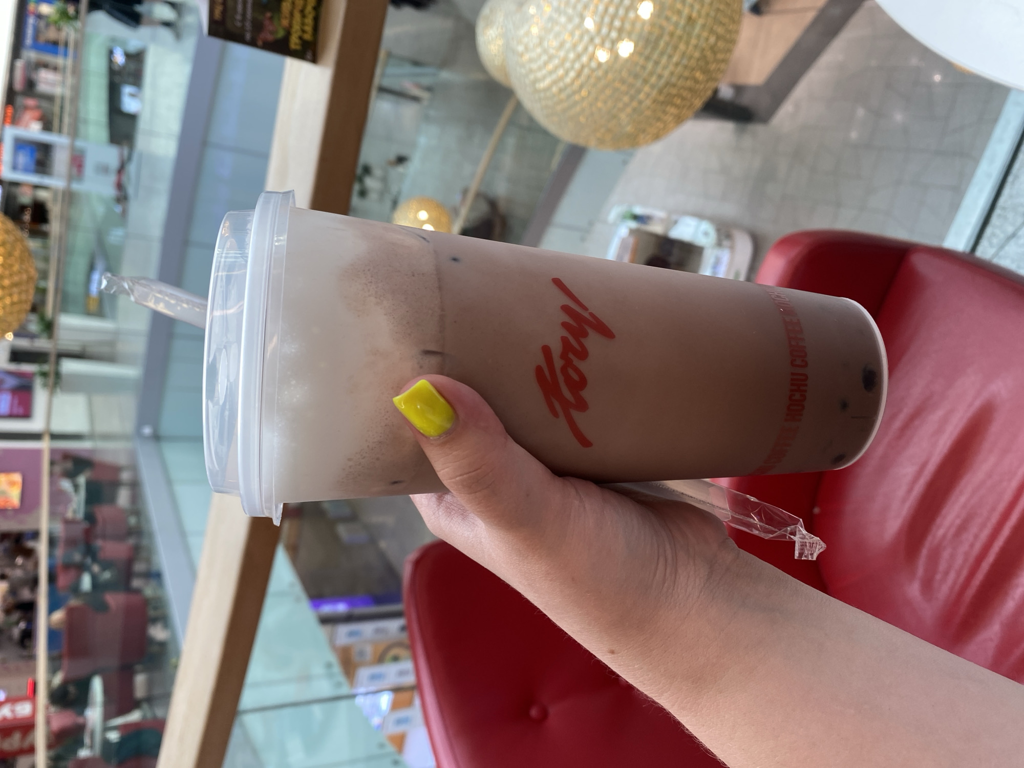 A person's hand with bright yellow nail polish holds a tall, clear plastic cup filled with a brown beverage and white foam at the top. The cup has red text on it that reads "Kory!" and below that, partially visible, "KOCHU COFFEE HOCHU". There are dark spheres, likely boba pearls, visible at the bottom of the cup. A plastic straw protrudes from the lid. The background suggests an indoor shopping mall with a balcony overlooking a lower level. Red seating is visible in the foreground, and decorative golden light fixtures are in the midground. The overall impression is a casual scene in a public space. The location is identified as Unknown, Russia.