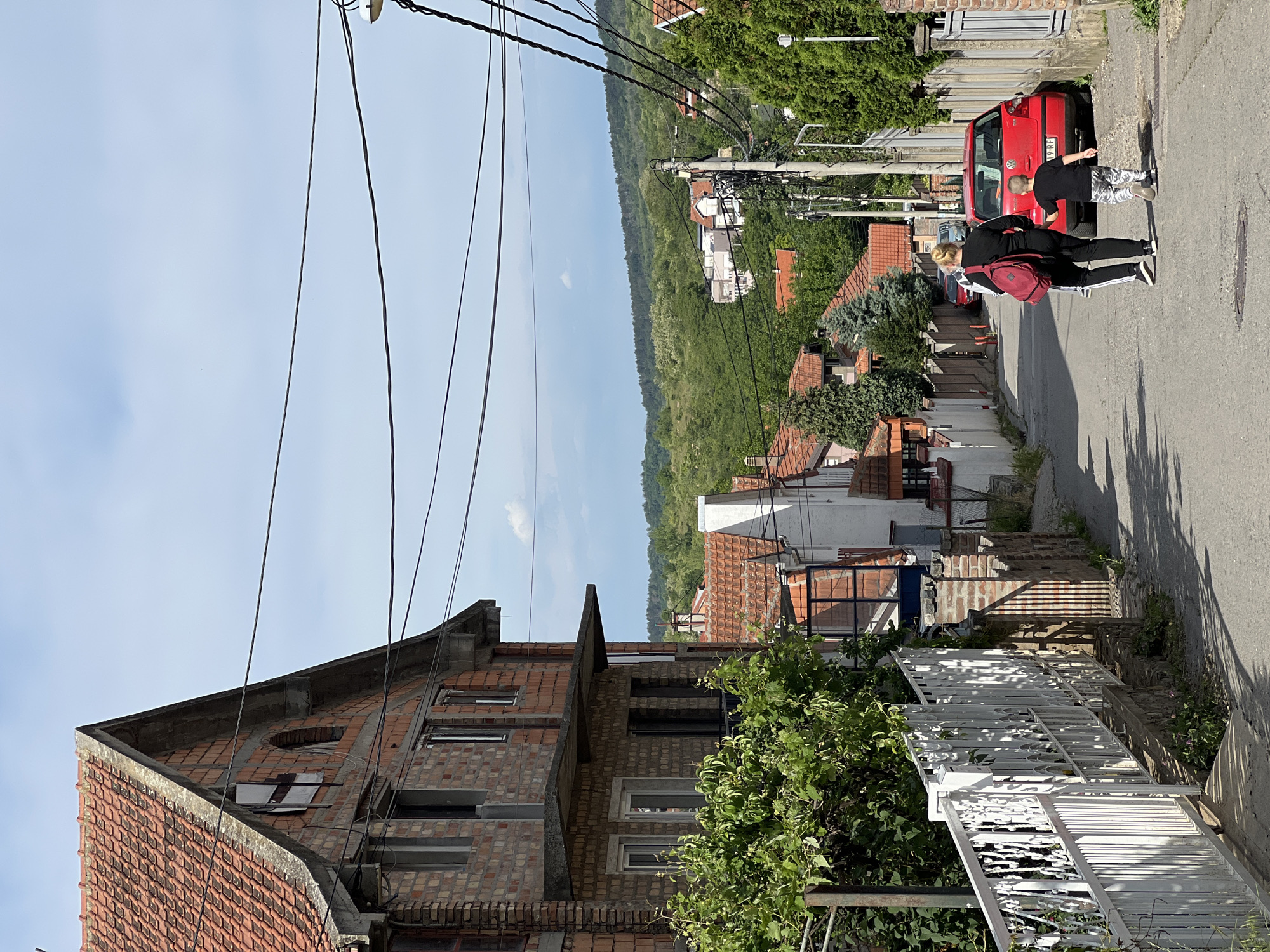 A daytime shot, slightly rotated, showcases a residential area of Palilula Urban Municipality, Serbia. A partially constructed brick building is on the left. Overhead power lines traverse the scene. A road stretches into the distance. On the road, a person with a red backpack is walking alongside a child toward a red car. Houses with red tile roofs are adjacent to the road. Green foliage is abundant in the background. A white fence is near the bottom of the frame.