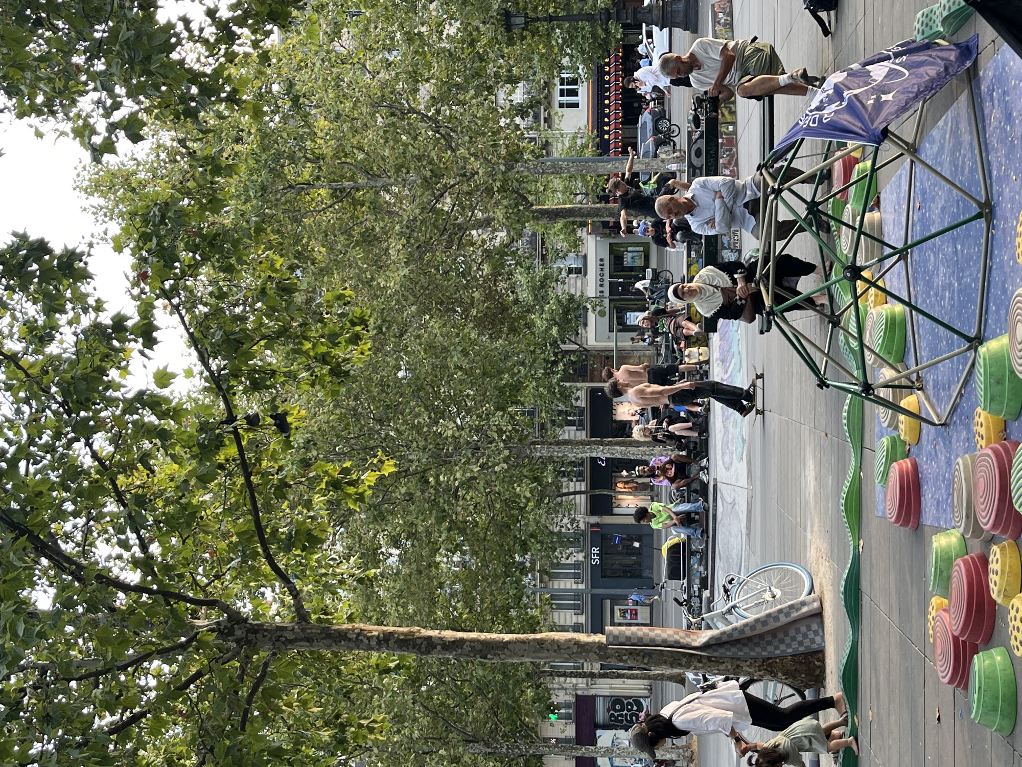 The image depicts an outdoor scene, likely in Paris, France, with a backdrop of buildings that appear to be shops and apartments. In the foreground, a colorful, low-profile children's play structure is visible, composed of a geodesic dome frame with a blue mat on the ground, surrounded by a series of green, yellow, and red semi-spherical stepping stones arranged in a wavy pattern.
Several people are present in the midground. A woman in a white shirt and black pants, holding the hand of a young child in a light green dress, is walking on the paved area. A bicycle with a yellow bag on its handlebars is parked near a tree trunk covered by a checkered mat. Further back, a group of people are sitting on a long bench. One individual, wearing a green shirt, is seated on the bench. Another person, wearing a dark hijab and a black outfit, is also seated. A man in a light blue shirt and dark pants sits next to her with his arms crossed. To his right, a man in a white shirt and khaki shorts is holding a video camera.
In the background, several more individuals are visible. A young man in a black shirt is skateboarding, performing a trick. Other people are standing or sitting, some of whom appear to be spectators or participants in an informal gathering. The area appears to be a public square or park, with trees providing shade over the paved and grassy areas. The overall atmosphere suggests a casual, daytime urban setting with recreational activities taking place.
