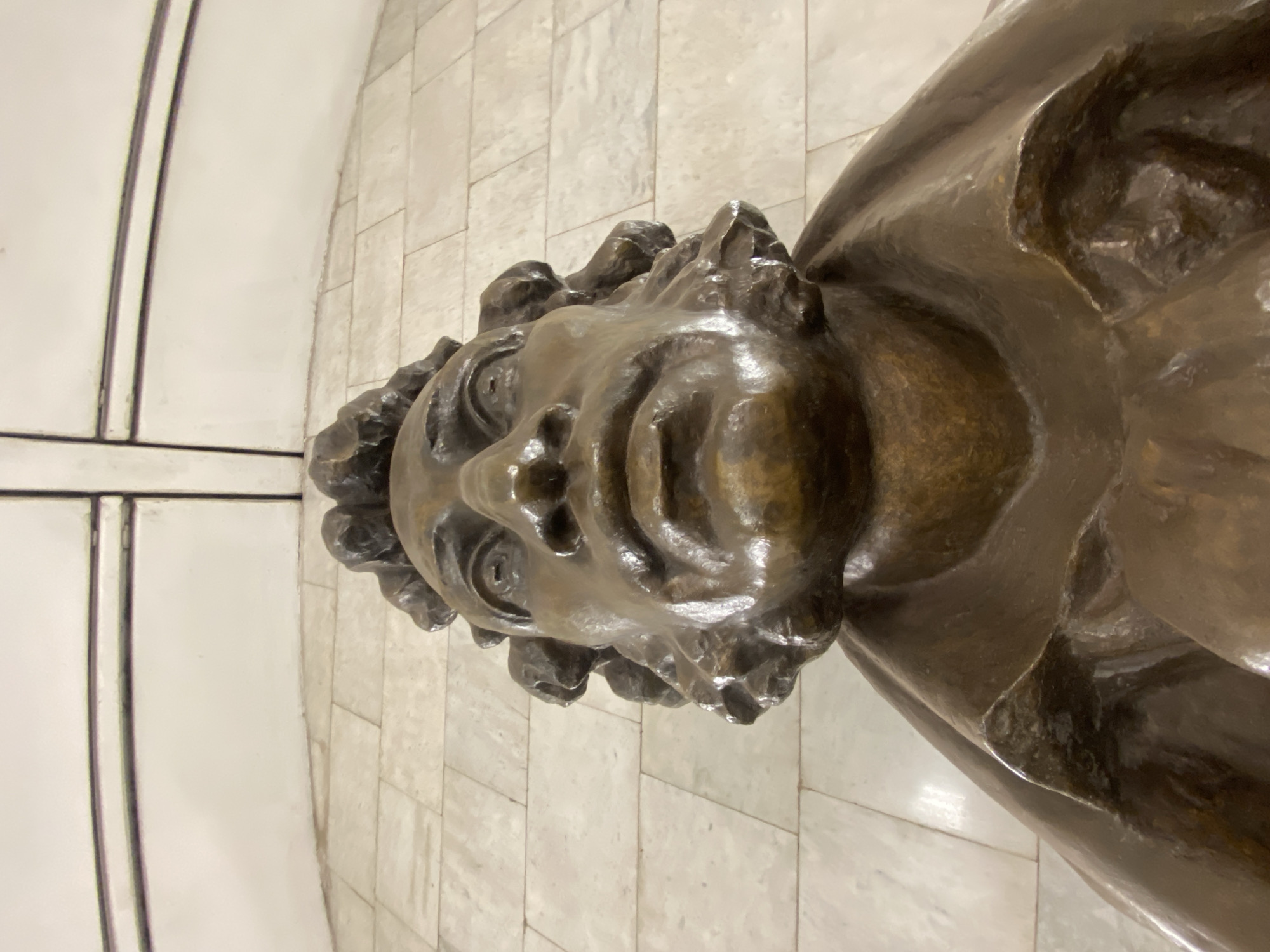 The image shows a close-up, high-angle view of a bronze bust, presumably a statue. The bust depicts a person with a distinctive, somewhat grotesque facial expression, featuring bulging eyes, a wide mouth, and a wrinkled forehead. The hair is curly and unkempt. The statue is positioned against a backdrop of light-colored, rectangular tiles. The location is Moscow, Russia. The statue is located in the Moscow Metro station Ploshchad Revolyutsii.