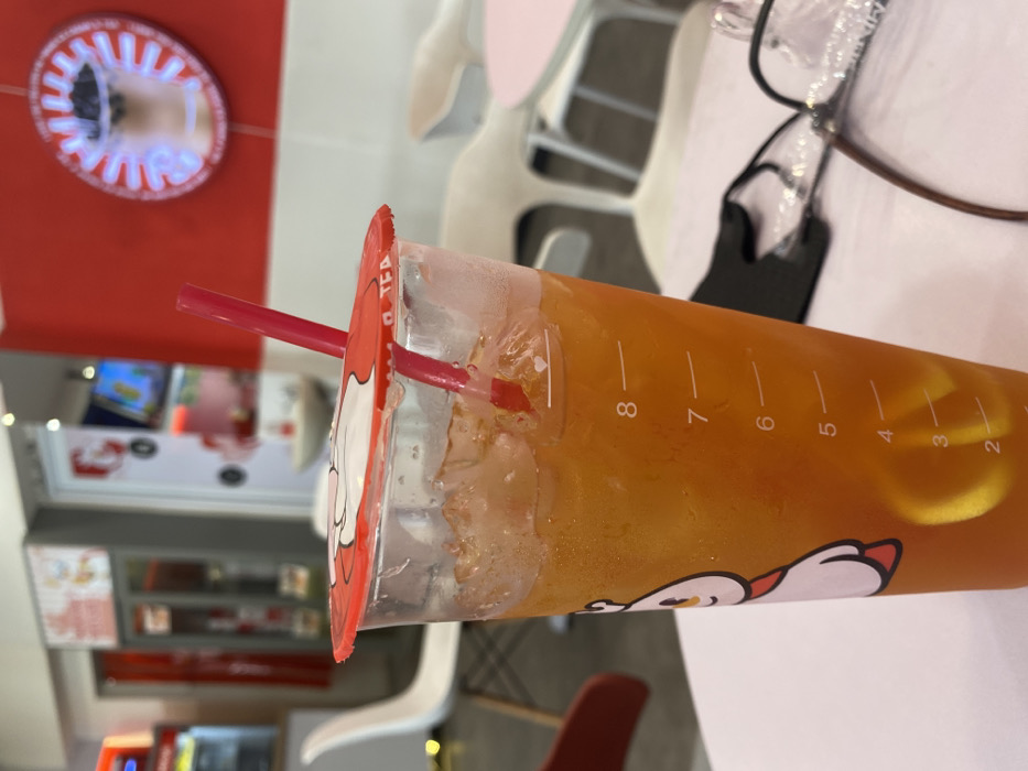 A close-up shot of a clear plastic cup filled with a bright orange liquid. The cup is labeled "O TEA" and has a red lid with a cartoon image of a snowman. A red straw sticks out of the top of the cup. The liquid in the cup is partially filled with ice, and the level is marked with measurements. The cup is sitting on a white table. The table is partially out of focus, but there are glasses, a black face mask, and a white chair in the background. The environment is a cafe or restaurant with red and white walls. There is a large red circle with a white interior on the wall behind the cup. The location is a shop in Kecamatan Piyungan, Indonesia, latitude: -7.837, longitude: 110.474.
