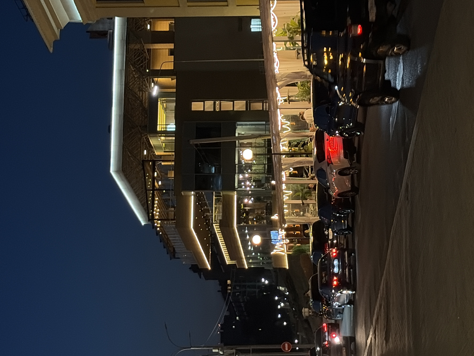 The image depicts a nighttime scene in Izhevsk, Russia, showing a multi-story building with illuminated architectural details. The building appears to house a restaurant or commercial establishment, with visible tables and chairs, some under canopies. Cars are parked along the side of the street, their tail lights visible. Streetlights and additional lighting from nearby buildings add to the ambient brightness. The sky is a dark blue, indicating it is late evening.