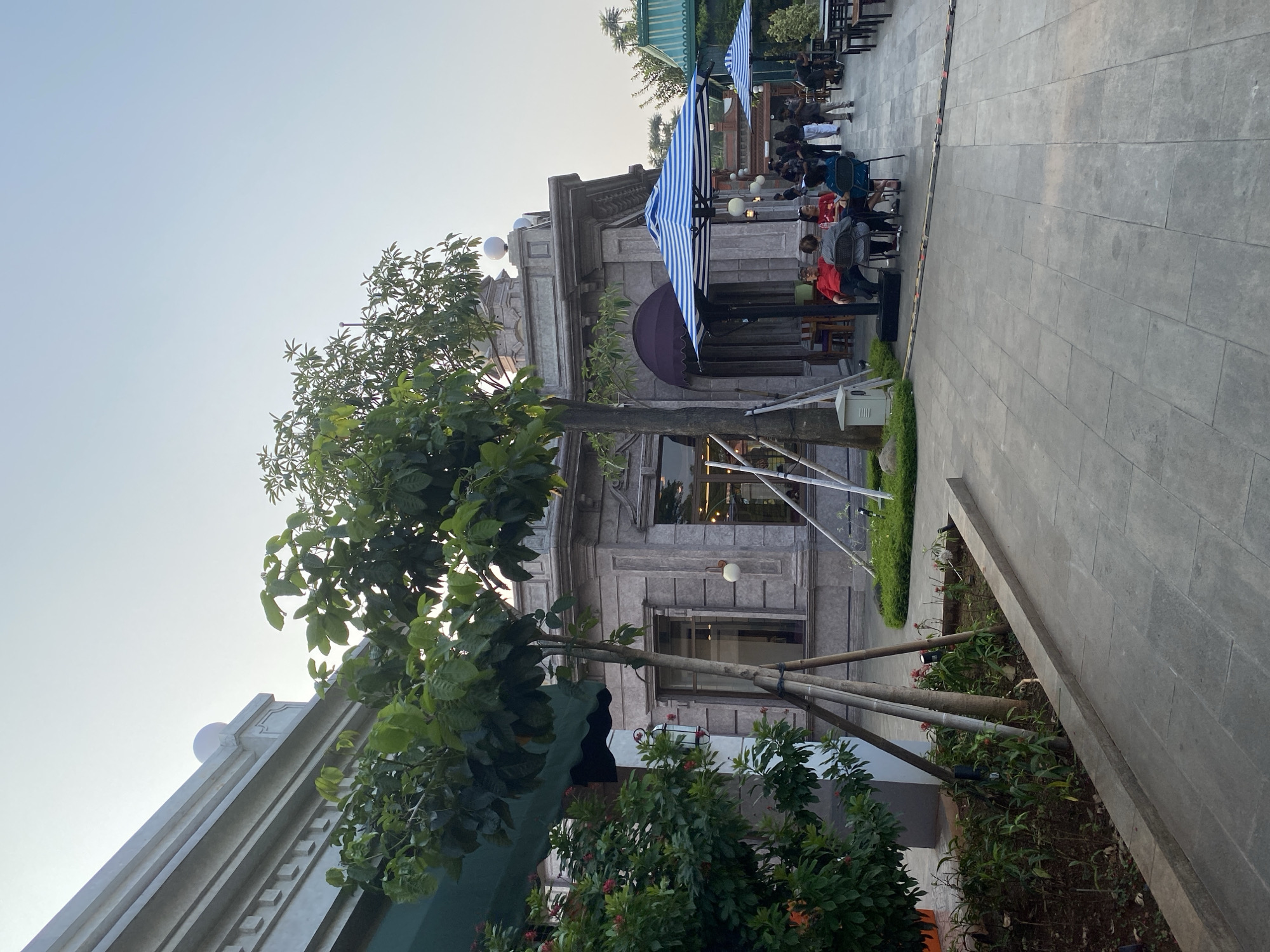The image depicts an outdoor seating area of a establishment, likely a cafe or restaurant, with people seated at tables. The scene is viewed from a slightly elevated angle, looking towards a building with classical architectural elements. In the foreground, a large tree with green foliage partially obscures the view.
Several people are visible, seated at tables in the outdoor area. Some are engaged in conversation, while others appear to be looking around. The people are casually dressed. Objects present include tables and chairs, and two prominent blue and white striped umbrellas, providing shade. The building behind the seating area has stone cladding and large windows. A paved walkway runs alongside the seating area and continues into the distance. There are also planters with greenery.
The time of day appears to be late afternoon or early evening, given the soft, diffused light and the clear sky. The weather seems pleasant. The architecture of the building and the presence of outdoor seating suggest an urban or semi-urban commercial setting. The overall mood is relaxed and social. No visible text is present in the image.