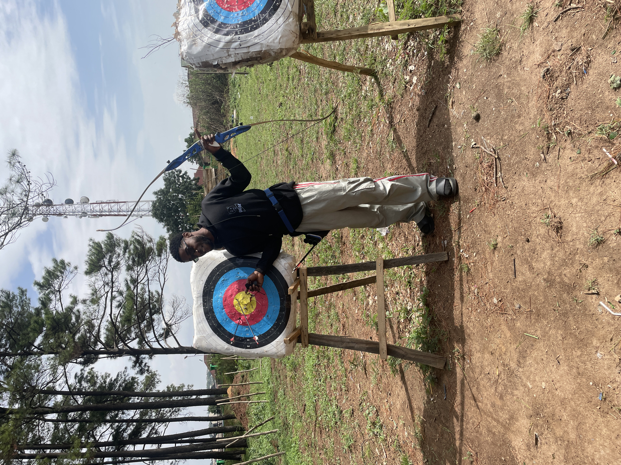 A man is standing outdoors next to an archery target in Jos, Nigeria. He is wearing a black jacket, grey pants, and black shoes. He is holding a blue bow, and an arrow is aimed towards the target. The archery target is circular and has concentric rings of different colors: red, yellow, blue, black, and white. There are several arrows already in the bullseye. The target is mounted on a wooden stand. Behind him and to the left are tall pine trees, and in the background is a telecommunications tower. The ground appears to be a mix of dirt and grass. There is another archery target and a stand behind the man on the right. The sky is partly cloudy.