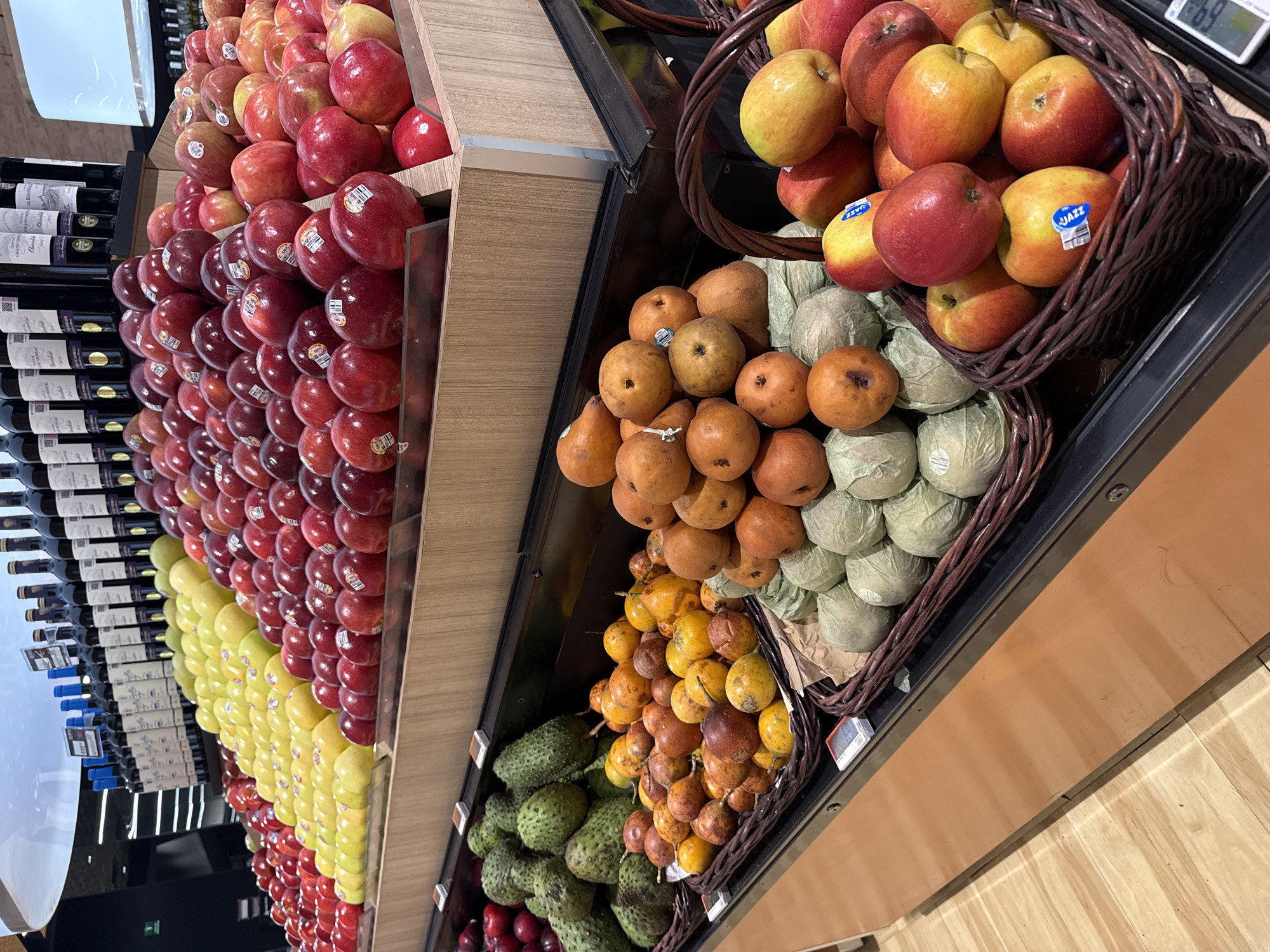 The image shows a grocery store display, likely inside a supermarket, featuring fresh produce and wine bottles. The location is plausibly in Puebla, Mexico, based on the types of fruits and the overall aesthetic.
On the left, stacked apples of different varieties (red, yellow, and a mix) form a large display. Behind the apples, wine bottles are arranged on shelves.
To the right, produce is arranged in baskets on shelves. From top to bottom, the baskets contain:
-A mix of red and yellow apples.
-Orange-skinned pears atop a layer of wrapped pale green fruits (possibly Asian pears or a type of melon).
-Yellow passion fruits.
-Green soursops at the very bottom, mixed with some red fruit.
The shelving and flooring are made of light-colored wood. The lighting is bright, suggesting an indoor supermarket environment. There are no people visible in the frame.