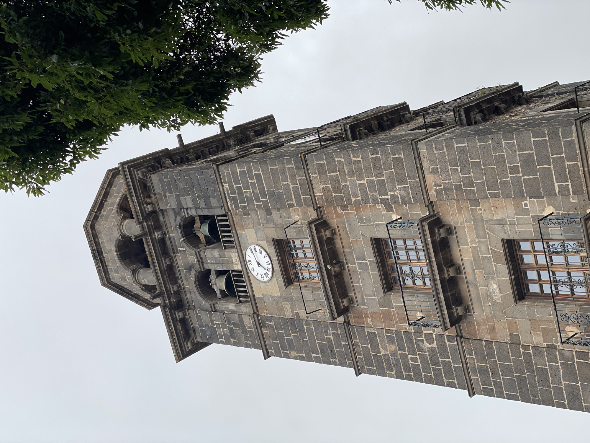 The image displays a tall, imposing stone bell tower, likely part of a church or historic building, viewed from a low angle against a uniformly overcast, light grey sky. The tower is constructed from dark, rectangular stone blocks, giving it a rugged and ancient appearance.
Towards the upper section, a circular clock face with Roman numerals from I to XII is clearly visible. The clock's hands indicate a time of approximately 1:50 or 2:50. Adjacent to the clock face and slightly above it, an arched opening reveals a large bell within, confirming its function as a bell tower. Another bell opening is partially visible further up.
Below the clock and on the lower visible sections of the tower, several rectangular windows are present. These windows are adorned with intricate, black wrought-iron grilles and feature wooden frames, adding decorative detail to the sturdy stone facade. A metal downspout runs vertically down one side of the tower, indicating rainwater drainage. The architecture also includes various decorative cornices and ledges that articulate the tower's levels.
In the upper left portion of the frame, the dark green foliage of a tree partially frames the tower, suggesting the presence of greenery nearby. The sky is completely clouded over, indicating an overcast daytime. No people or specific activities are depicted; the scene is static, focusing solely on the architectural structure.
The only visible text consists of the Roman numerals on the clock face. The architecture is consistent with historical European structures, and the provided context places this location in San Cristóbal De La Laguna, Spain.
