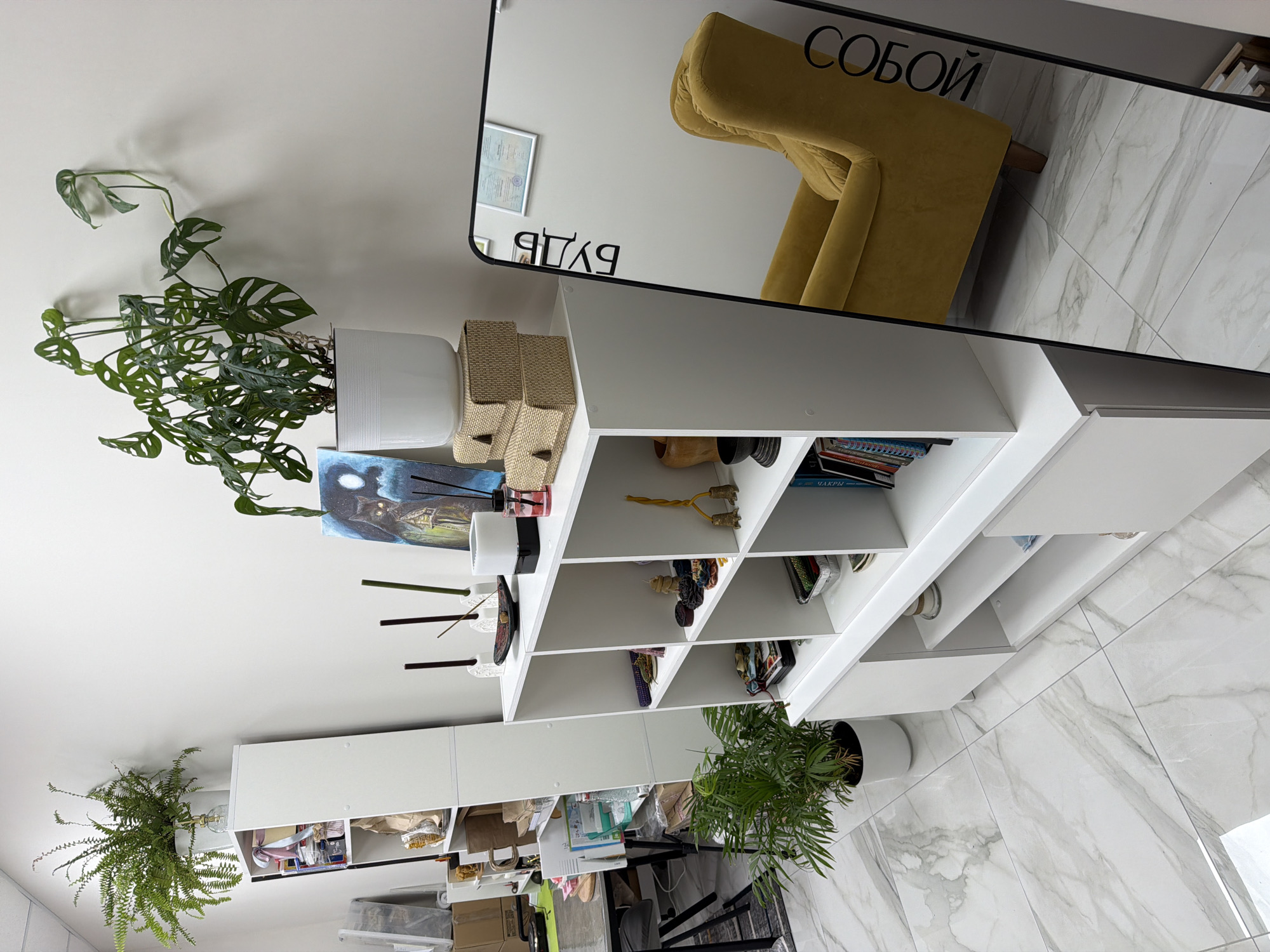 The image depicts an indoor scene, likely a room or studio space, with clean, modern white shelving units and a white tiled floor. Several potted plants are visible, including a large fern on top of a taller shelf unit on the left and a smaller palm-like plant on the floor in front of a lower white unit. On the shelves, various decorative objects are displayed, such as candles, a painting of a cat under a moon, small statues, and stacks of woven boxes. A large standing mirror with a black frame is positioned on the right side of the image, reflecting a yellow armchair and part of the room. Text in Cyrillic is visible on the mirror, reading "БУДЬ СОБОЙ" which translates to "BE YOURSELF". The overall impression is of a well-organized and aesthetically pleasing space. There is no indication of the city of Petropavlovsk-Kamchatsky, Russia, or any specific geographical markers in the image.