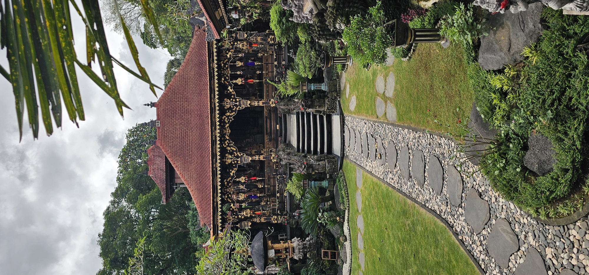 The image depicts a Balinese temple in Ubud District, Indonesia. A stone pathway leads to the temple entrance with stairs. The temple has an ornate, multi-tiered roof and intricate carvings. Lush vegetation surrounds the temple and pathway.