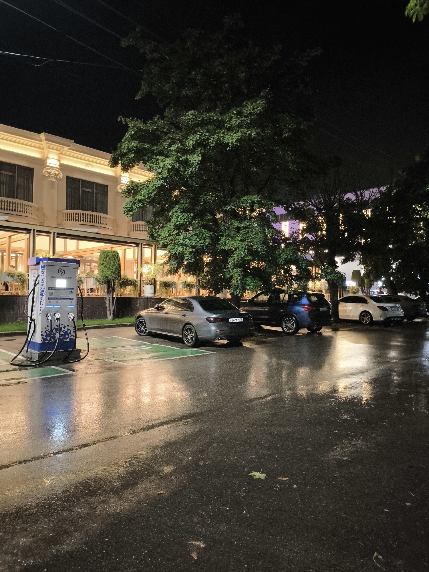 The image depicts a night scene on a wet asphalt street in Yessentuki, Russia, likely in front of a building with classical architectural elements and illuminated windows, possibly a restaurant or hotel. A prominent electric vehicle charging station, blue and white with multiple charging ports, stands on a designated green parking space. Parked to the right of the charging station is a silver sedan. Further down the street, other vehicles are visible, including a dark SUV and a white sedan. Lush green trees with dense foliage are scattered throughout the scene, their leaves reflecting the ambient light. The ground is wet and reflective, indicating recent rain or ongoing precipitation, with streaks of light from the building and streetlights creating a shimmering effect. The overall atmosphere is dark and somewhat moody, with artificial lighting casting warm glows on the wet surfaces and illuminating the building's facade. There are no visible people interacting with the charging station or vehicles in the immediate vicinity, though figures can be seen through the illuminated windows of the building. The sky is dark, suggesting it is nighttime.