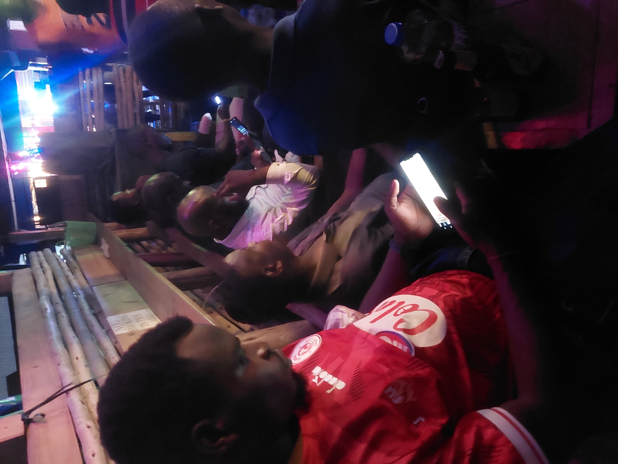 This indoor image captures a nighttime gathering of several people in a dimly lit, casual setting, likely in Dar es Salaam, Tanzania. The scene appears to be a social venue with rustic wooden structures, possibly benches or platforms, on which people are seated or reclining.
Multiple individuals, predominantly men, are visible throughout the frame. Many are interacting with their mobile phones, whose bright screens cast light onto their faces and hands. In the foreground, a man lies on his back, wearing a red football jersey featuring the "diadora" brand logo and a club crest, consistent with Simba SC, a popular team in Tanzania. Other people are seated or standing further back, also appearing relaxed and engaged in conversation or their phones.
The environment is characterized by dramatic lighting, with a strong, bright blue and white light source emanating from the left side of the frame, creating flares and illuminating parts of the scene. This artificial light contrasts sharply with the predominantly dark background and foreground areas. The wooden construction elements, along with the casual attire and use of mobile phones, suggest a laid-back, informal gathering or social event. The overall atmosphere appears relaxed and social. No other significant text apart from the jersey branding is clearly discernible.
