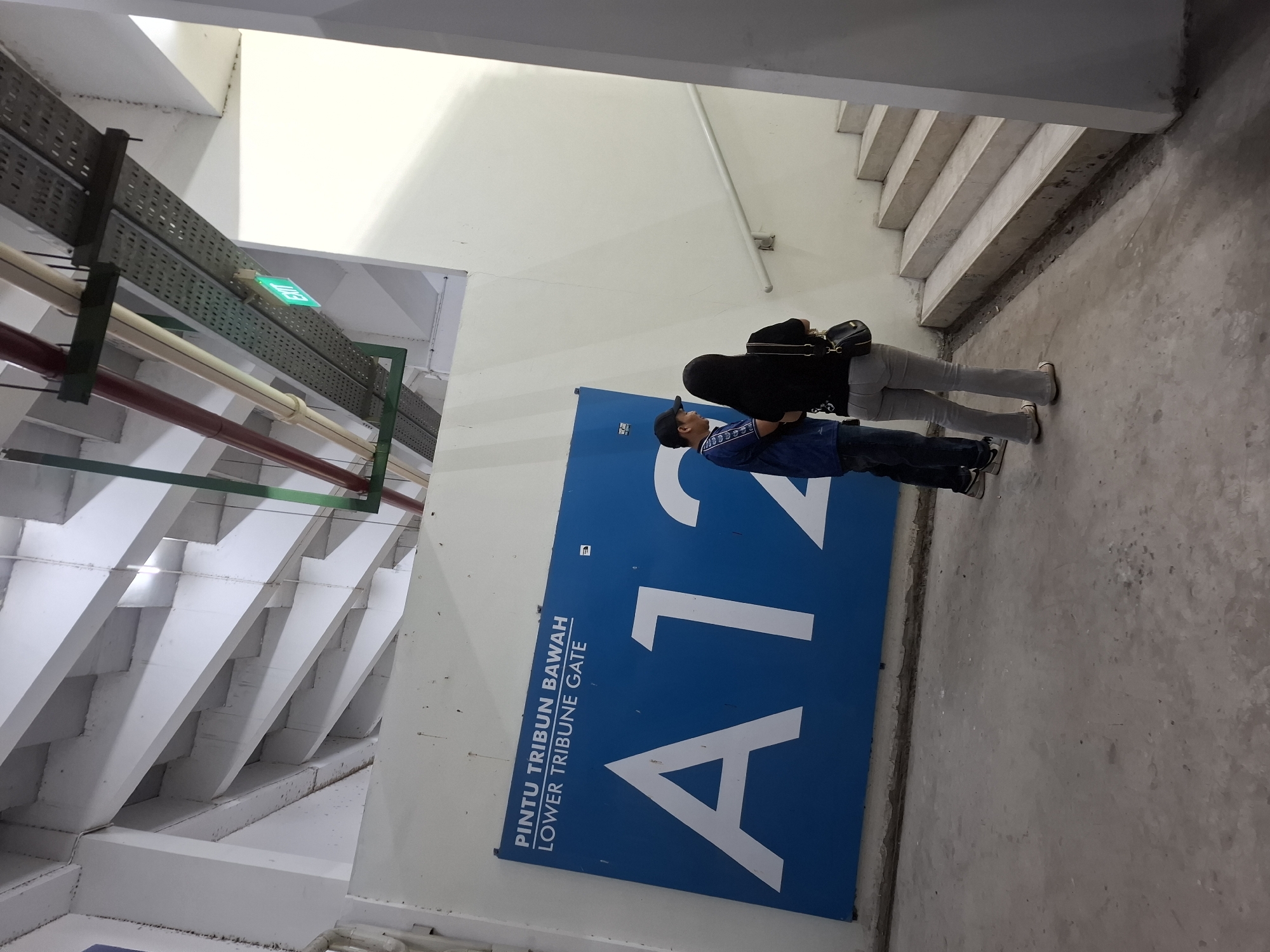 In the city of Perumda Deket, Indonesia, two individuals, a male and a female, are standing next to a large blue sign that reads "PINTU TRIBUN BAWAH LOWER TRIBUNE GATE A12". The man, wearing a blue shirt and a cap, is facing the sign and appears to be looking at it. The woman, dressed in a black top and grey flared pants, is facing away from the sign and looking up towards a staircase. She is carrying a black shoulder bag.
The environment is an indoor walkway with a concrete floor and a white wall. Above, there are exposed pipes and conduits running along the ceiling, with a green "EXIT" sign visible. The staircase on the right is made of concrete. The lighting appears to be artificial, casting shadows. The overall scene suggests an entrance or passage within a stadium or a similar large public venue.