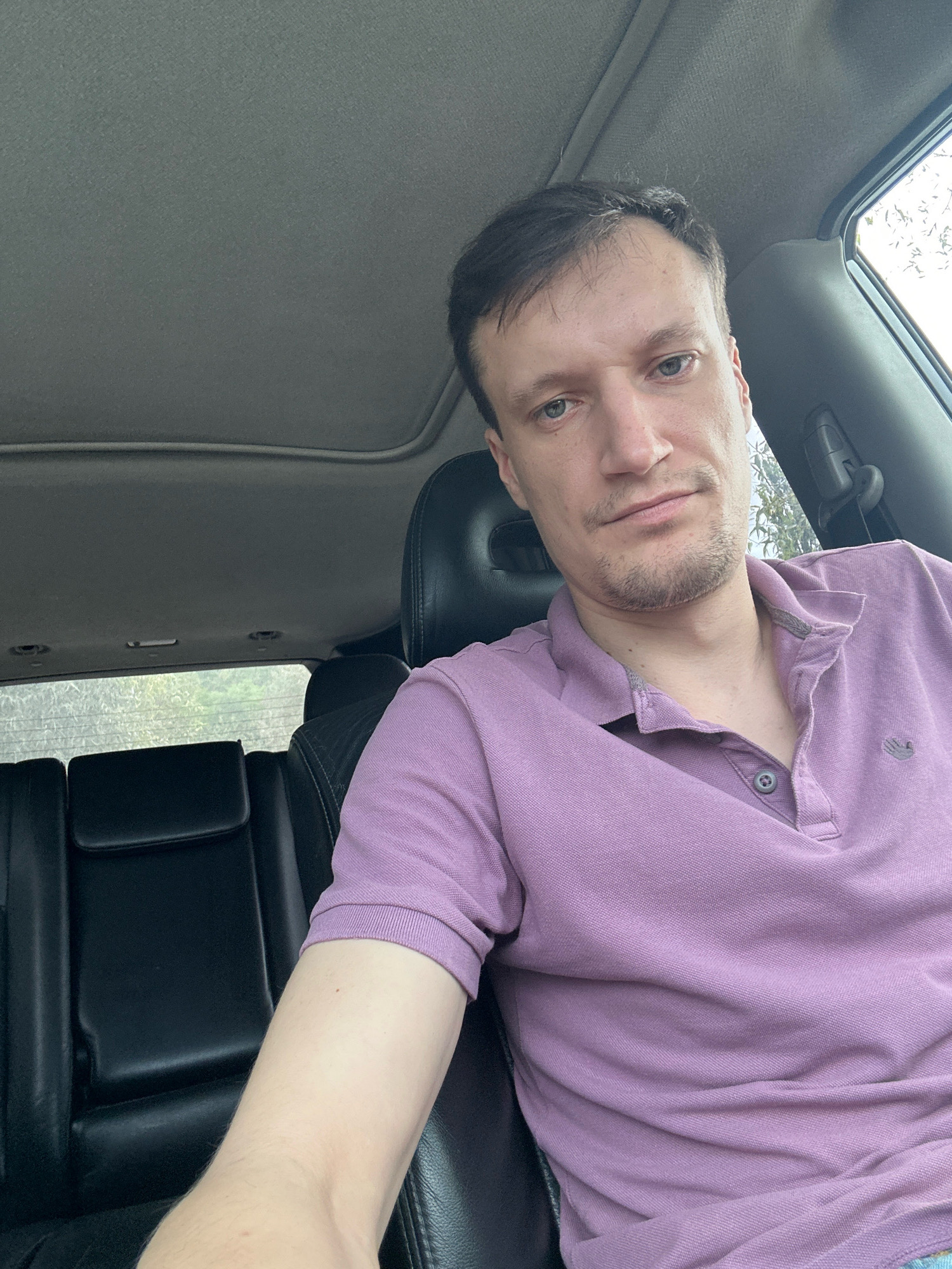A medium shot shows a man sitting in the driver's seat of a vehicle, looking at the camera. The man has light skin, short brown hair, and is wearing a light purple polo shirt. He is in the front passenger seat of a car, with the back seats visible behind him. The interior of the car is dark, and there is a sunroof overhead. The man appears to be in Makiv, Ukraine.