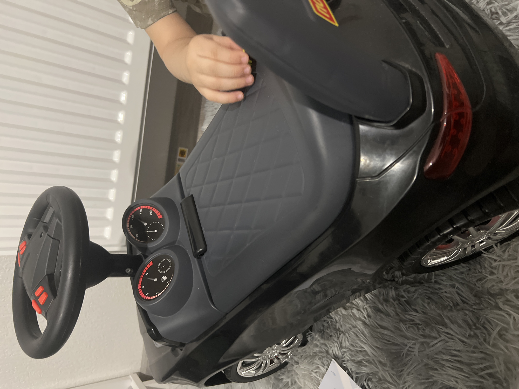 The image shows a close-up of a black ride-on toy car, possibly for a child. The car has a steering wheel with red buttons, two dashboard gauges, and a textured seat. A small child's hand is visible, touching the seat. The car is positioned on a fluffy gray rug. In the background, there appears to be a white radiator or heating unit against a light-colored wall. There is no discernible information to indicate the location is Yekaterinburg, Russia.