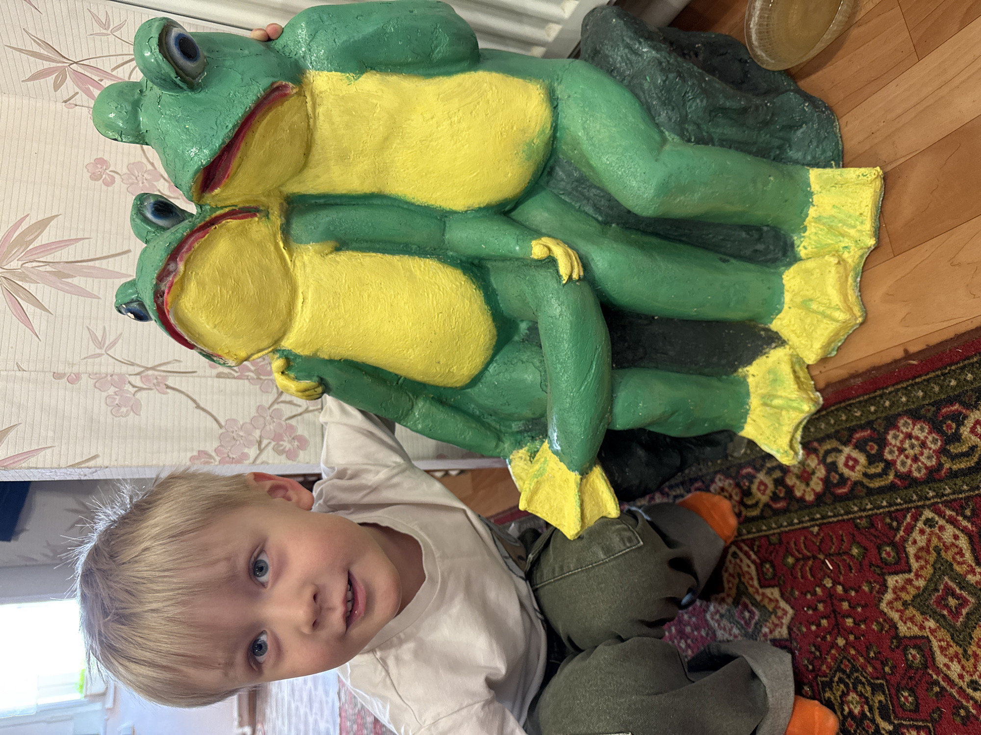 A young child with blonde hair and blue eyes, wearing a light-colored long-sleeved shirt and green pants with orange socks, is looking up at the camera. He is sitting on a patterned rug with red, black, and gold colors. Next to him is a large, painted statue of two frogs stacked on top of each other. The frogs are primarily green with yellow bellies and feet. The statue is placed on a dark, possibly rocky base. In the background, there is a wall with a floral wallpaper pattern and a wooden floor. There is also a partial view of a radiator. The scene appears to be indoors.