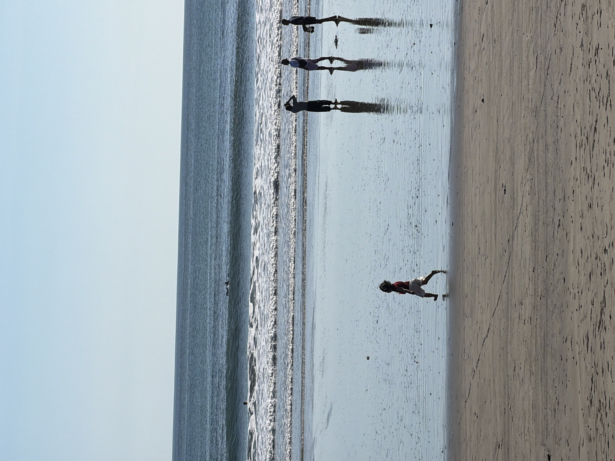 A group of people are captured in silhouette on a beach in Kuta, Indonesia, their figures elongated by the sun's glare on the wet sand. The image, oriented vertically, shows the ocean on the left with gentle waves lapping at the shore, and the sandy beach extending to the right.
In the water, several figures are seen standing, their shadows stretching across the reflective surface. Further up the beach, a child in a red top and white shorts is in mid-stride, appearing to run or jump. The scene suggests a bright, sunny day, likely in the late afternoon or early morning given the long shadows. The overall mood is tranquil, with the natural beauty of the beach setting taking center stage. There is no visible text in the image.