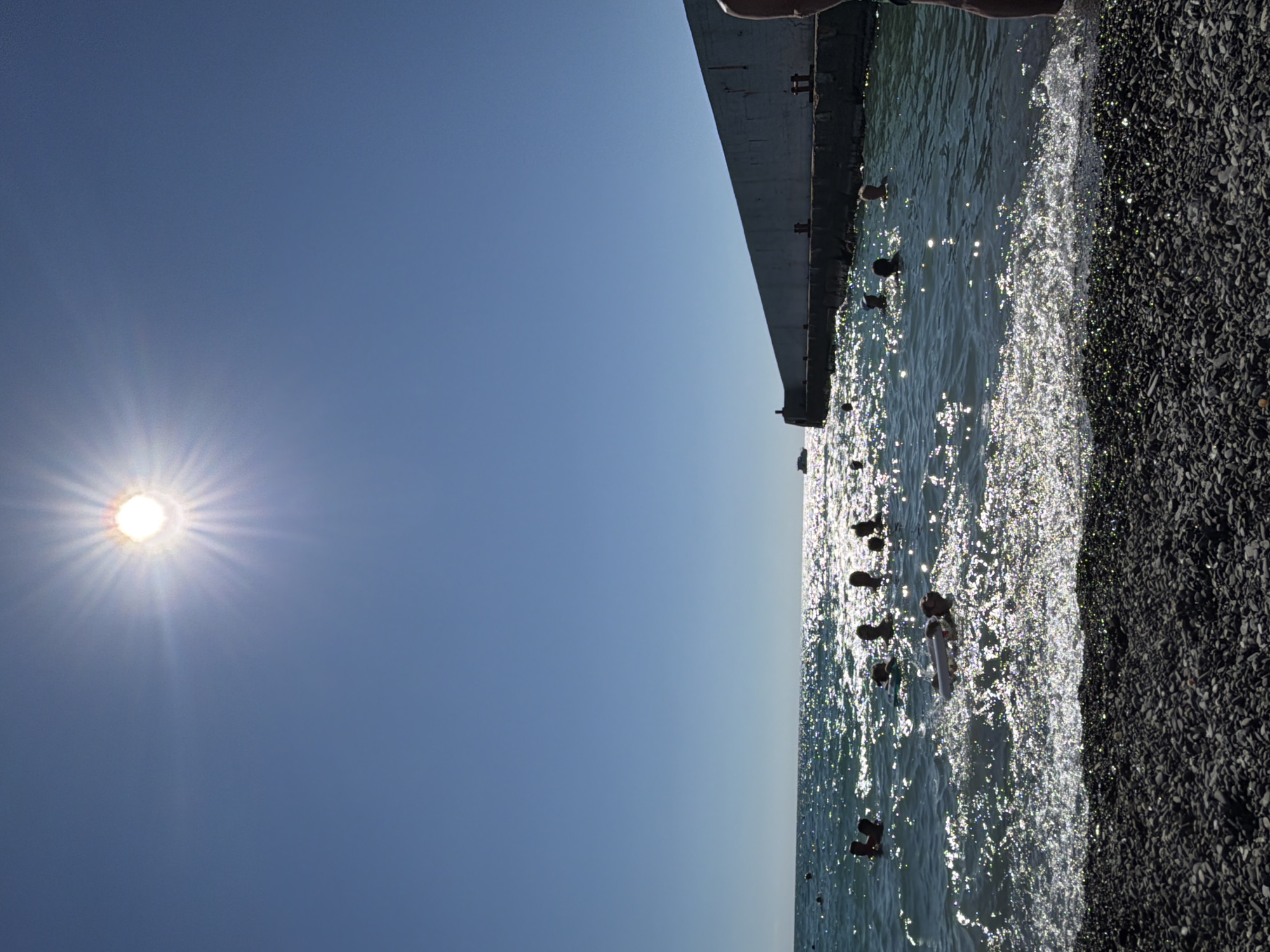 A sunny day at a pebble beach in Sochi, Russia, features several people swimming and playing in the sea. The sun is high in the sky, casting bright reflections on the water. A long, concrete breakwater stretches into the sea, with a small boat visible in the distance. The water is clear and a deep blue, with gentle waves lapping at the shore. The beach is covered in dark grey pebbles. A person's legs and feet are visible in the lower right corner of the frame, suggesting they are standing on the shore.