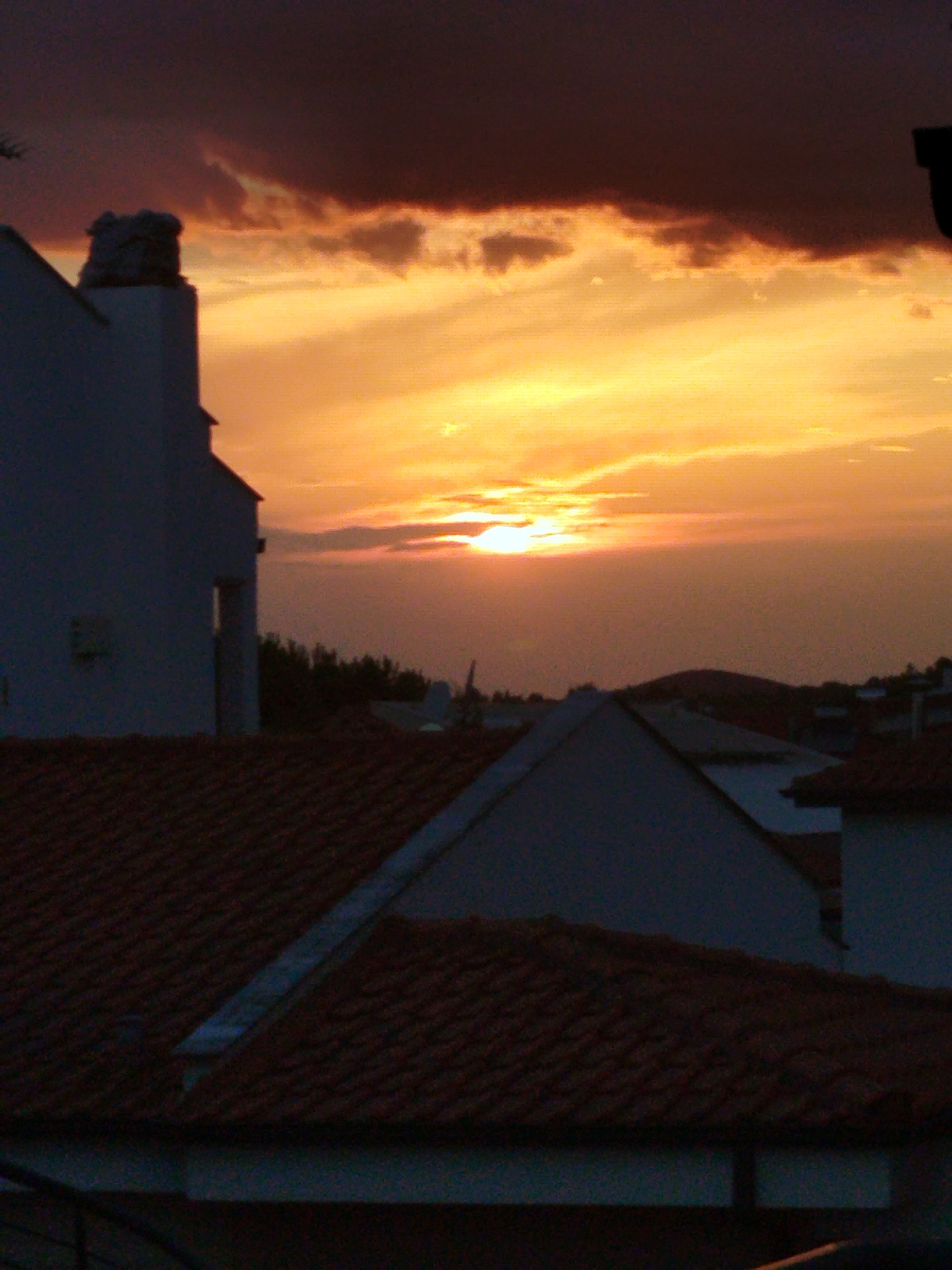 The image depicts a sunset over a residential area in Sithonia Municipal Unit, Greece. The sky is filled with a gradient of colors, from deep orange and yellow near the horizon to dark purple at the top. Clouds are scattered across the sky, with some catching the light and appearing bright. In the foreground, the terracotta tiled roofs of several houses are visible, with their characteristic dark red color. Silhouettes of white buildings with chimneys can be seen emerging from behind the rooftops. A few trees are visible in the middle ground, adding a touch of green to the scene. The overall impression is one of a tranquil evening in a coastal town.