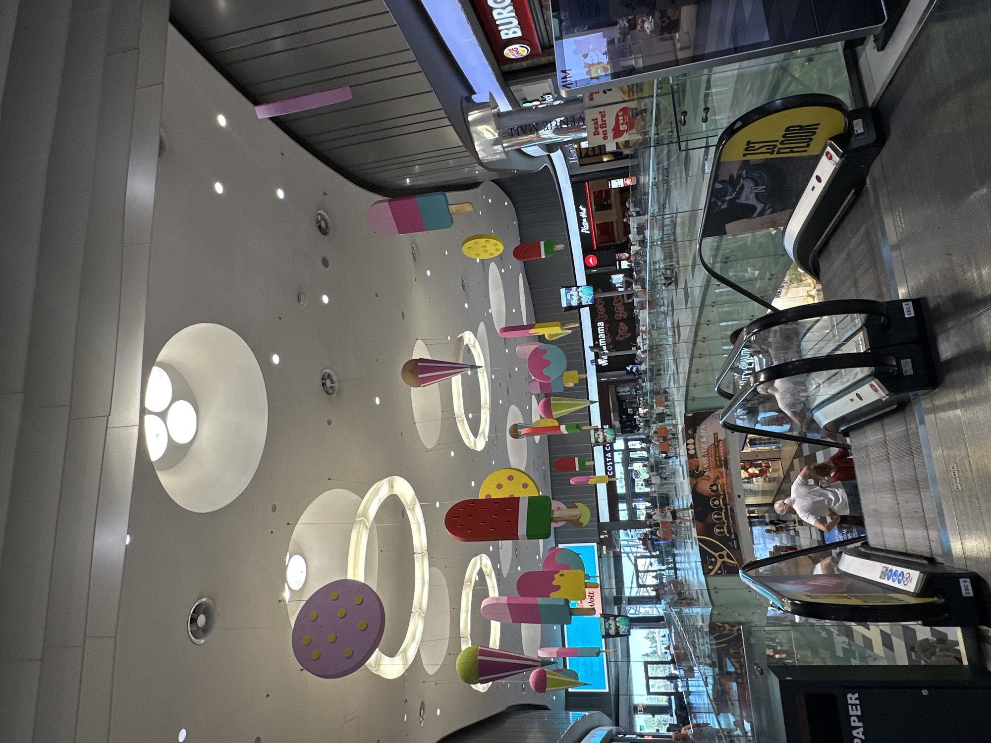 The image depicts the interior of a modern, multi-level shopping mall in Paphos Municipality, Cyprus. The atrium is characterized by a high ceiling with circular light fixtures and decorative hanging elements resembling oversized ice cream cones and cookies, suggesting a whimsical theme. Escalators are visible in the foreground, with several people using them. The mall appears to be populated, with shoppers browsing at various retail outlets visible in the background, including Burger King and Pizza Hut. The architecture includes glass railings and metallic accents, contributing to a contemporary aesthetic. The overall environment suggests a bustling commercial space designed for public recreation and shopping.