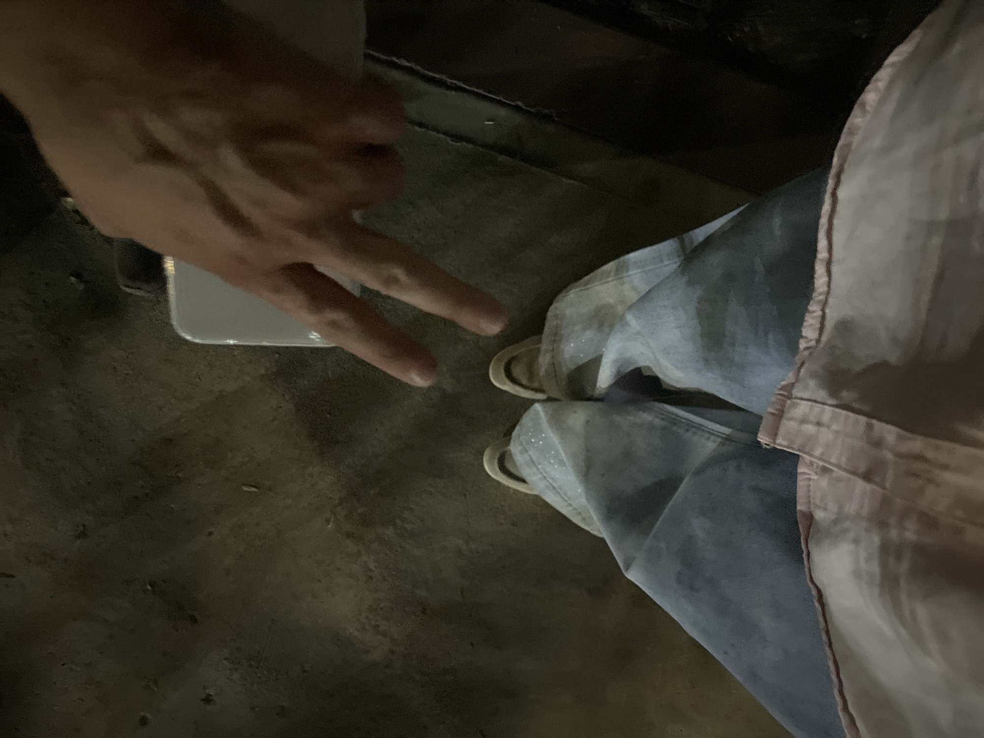 The image depicts a close-up, aerial view of a person's lower body, focusing on their legs and feet, positioned on a paved surface that appears to be a sidewalk or street. The person is wearing light-colored, possibly denim, wide-leg pants with subtle shimmering details, and light-colored sneakers. A hand, belonging to another individual, enters the frame from the top right, with two fingers extended in a V-shape, pointing towards the person's shoes. The hand is holding a white rectangular object, possibly a smartphone, which is partially obscured. The ground is rough and textured, with visible imperfections and debris. The lighting suggests it might be evening or nighttime, with artificial light sources casting shadows and creating a slightly muted color palette. The context of Tashkent, Uzbekistan, is not visually discernible from the image itself.