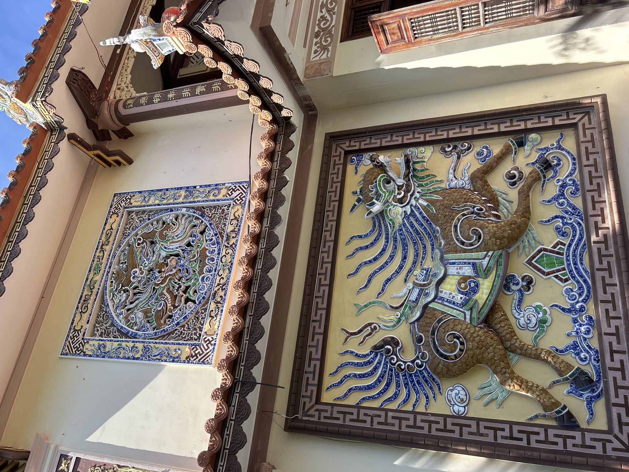 The image shows the exterior of a building with ornate decorative art, likely located in Tay Nha Trang Ward, Vietnam. The building has a light yellow painted exterior. The walls are adorned with two decorative art pieces. One is a square tile mosaic panel with an intricate circular design in blue, white, green, and brown colors. The other is a rectangular raised tile panel depicting a mythical creature resembling a horse or dragon, with a mosaic style. The tiles are in shades of brown, blue, white, and green. The creature is in a dynamic pose. Both panels are framed with decorative borders. The building's roof has a terracotta tile trim with a stylized pattern, suggesting traditional Vietnamese architecture. There is also a wooden balcony visible on the upper part of the building. Sunlight creates shadows on the walls, highlighting the textures and details of the architectural elements.
