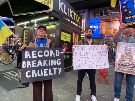 Captured in New York City, United States, the photograph features a group of three individuals engaged in a protest. They are positioned in front of a "KLIKTIX" ticket booth, amidst the bustling Times Square environment. The central figure holds a sign proclaiming, "TODAY, RUSSIA LAUNCHED ITS LARGEST ATTACK ON UKRAINE EVER." To his left, an elderly man stands holding a sign that reads "RECORD-BREAKING CRUELTY" and to his right, a man holds a sign showing a picture of a man, likely Vladimir Putin, with the caption "A WAR CRIMINAL." The Ukrainian flag is seen in the scene, reinforcing the thematic focus of the protest, likely concerning the conflict in Ukraine. Additional individuals and structures visible in the background contribute to the sense of the location being a crowded public space.