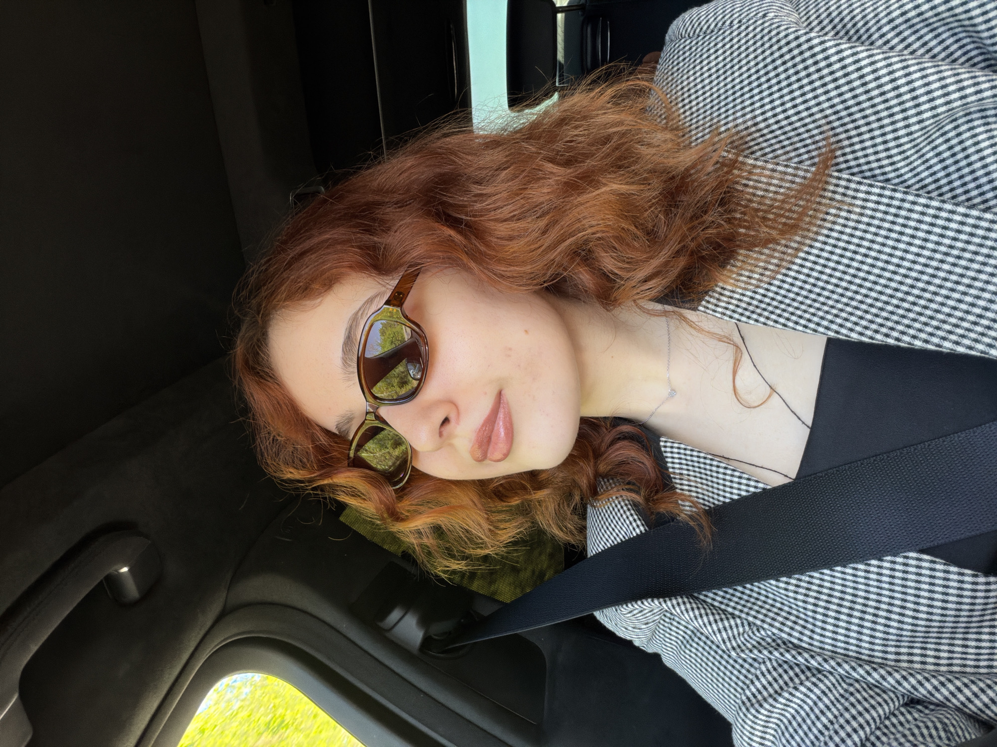 A young person, likely female, is seated in a car, looking towards the camera with a subtle, relaxed smile. She has reddish-brown, wavy hair and is wearing brown-framed sunglasses that reflect bright green foliage and a clear sky, indicating sunny weather. Her lips are full and appear to have a soft gloss. A thin silver necklace with a small pendant is visible on her neck.
She is dressed in a black and white small-checked jacket, resembling gingham or houndstooth, and a black seatbelt is securely fastened across her chest and shoulder. The car's interior is dark, with elements like the ceiling lining, a grab handle, and door frame visible. Through the car window, vibrant green trees or bushes can be seen, suggesting an outdoor, possibly rural or suburban, setting. The overall mood is calm and casual, captured during daylight hours in Strasbourg, France. No visible text is present in the image.