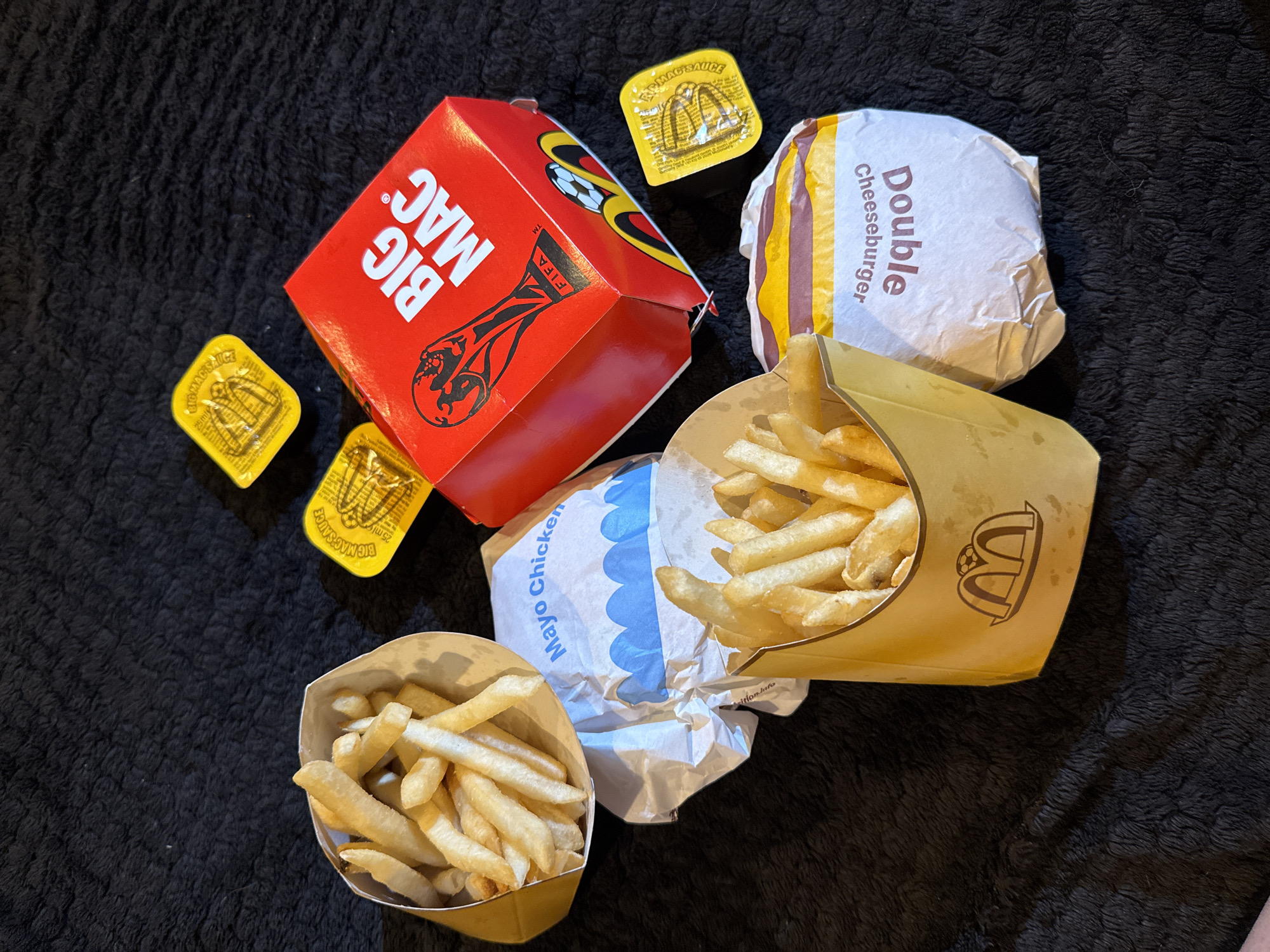 The image displays a spread of fast food items from McDonald's, arranged on a black, textured surface, likely a blanket or rug. The items include two servings of french fries in their signature brown paper containers, one wrapped "Mayo Chicken" burger, one wrapped "Double Cheeseburger", and a red "Big Mac" box. There are also three small, yellow sauce packets, one of which is clearly labeled "Big Mac Sauce". The "Big Mac" box features a FIFA World Cup trophy graphic. The setting is indoors, with no visible windows or external environment. There are no people or animals present in the image. No specific location within Great Yarmouth, United Kingdom, can be identified from the visual information provided.