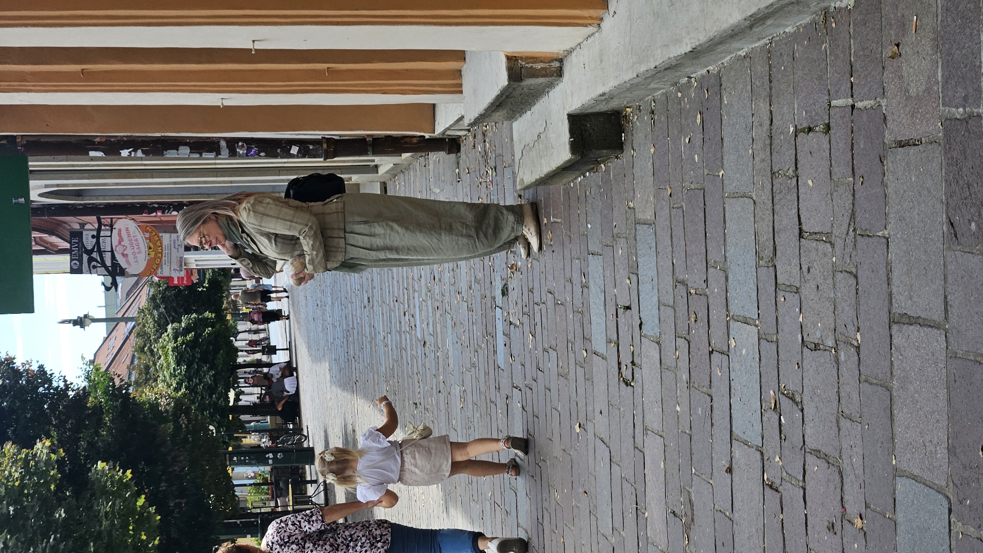 The image depicts a cobblestone street scene in the city of Prešov, Slovakia, likely on a moderately overcast day. In the foreground, a woman with blond hair, wearing a light-colored top and a tan skirt, is walking away from the camera, holding the hand of a young girl with blonde pigtails who is also walking away. The girl is wearing a white t-shirt and a light-colored skirt. To the right of the foreground figures, a woman with graying blond hair, wearing glasses, a light-colored checkered jacket, and loose-fitting light green pants, stands facing left. She is holding a light-colored object in her hands and appears to be looking towards the left side of the frame. Behind these individuals, the street continues with more people walking and sitting. The buildings lining the street show architectural details characteristic of European cities, with some featuring arches and striped facades. Signage is visible on the buildings, suggesting shops or businesses. Trees are planted along the sides of the street, providing shade. The cobblestone pavement is aged and shows some signs of wear. The overall atmosphere is that of a public urban space with pedestrian activity.