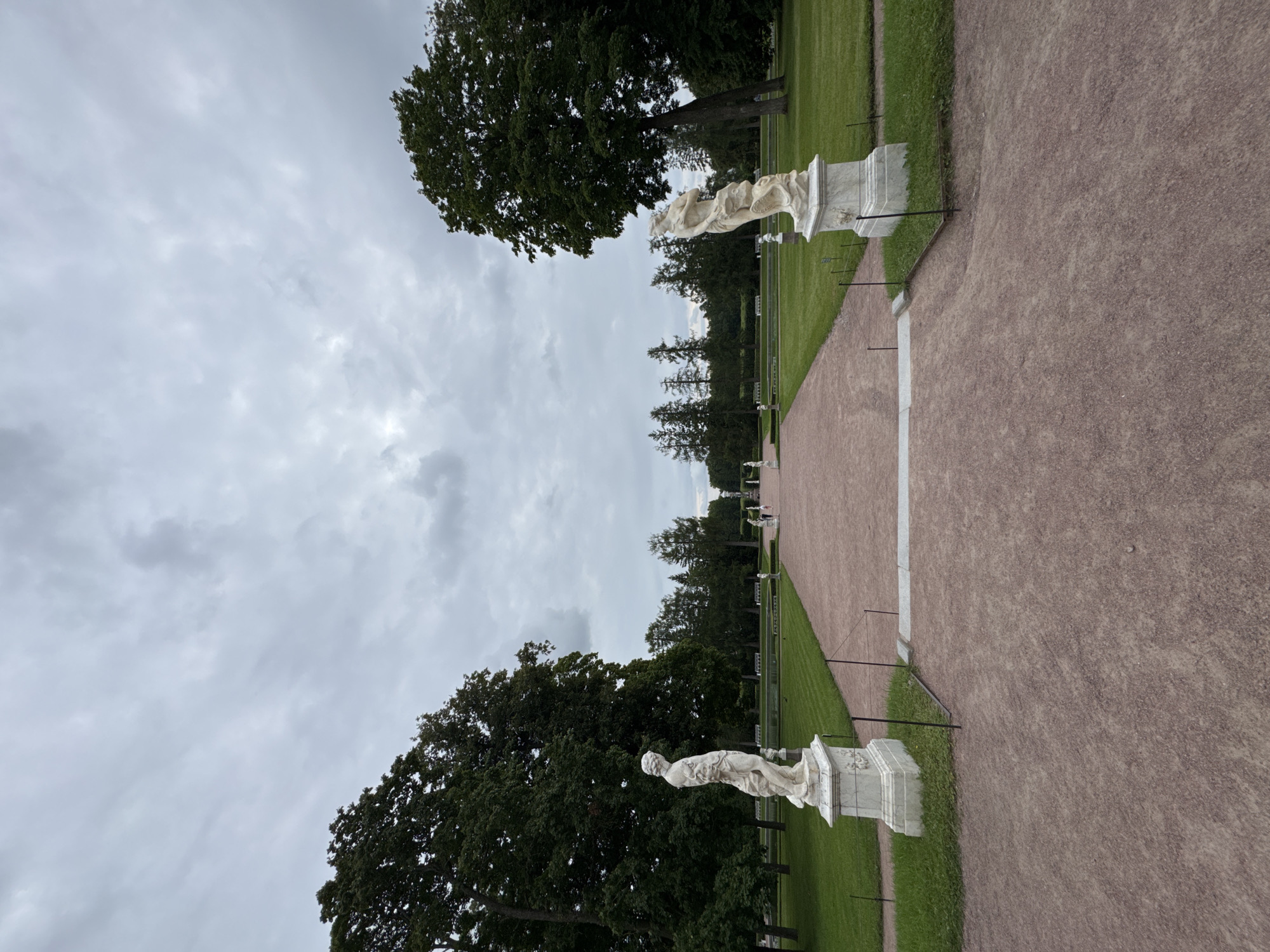 This image depicts a frontal view of a pathway in a park, with two classical marble statues on either side of the path. The pathway, made of reddish gravel, leads into the distance where more statues and a large pond or canal are visible, flanked by manicured lawns and trees. The sky is overcast with gray clouds. The statues are life-sized and depict human figures in classical drapery, standing on white marble pedestals. The park appears to be well-maintained, with neat green lawns and orderly rows of trees in the background. This scene is characteristic of the Catherine Park in Pushkin, Russia, specifically the area around the Grand Cascade.