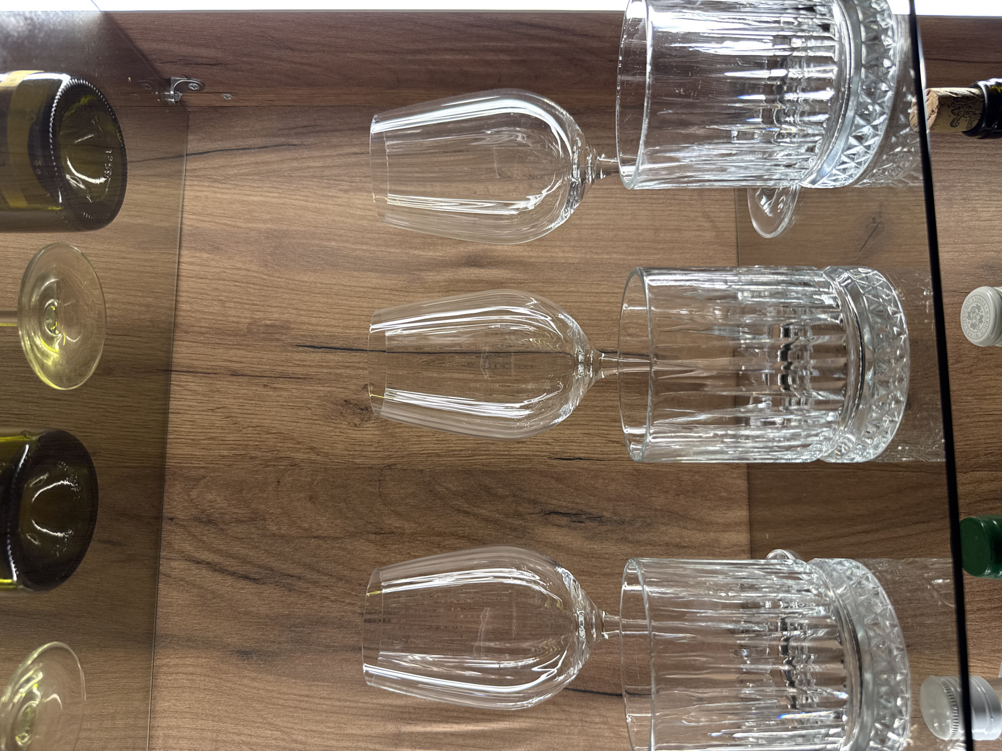 The image displays a well-organized cabinet with wooden paneling and glass shelves. On the top shelf, partially visible are three wine bottles and two wine glasses, their bases resting on the shelf. The middle shelf holds three stemmed wine glasses, each positioned above a distinct pattern of glassware. Below these, on the lowest visible glass shelf, are two cut-glass tumblers, likely for whiskey or spirits, with intricate patterned bases. Reflecting the light, these tumblers sit on the polished surface of the shelf. The bottom of the frame shows the edge of another glass shelf with various bottle caps and a corked bottle, hinting at a collection of beverages. The overall impression is one of a curated display, possibly in a home bar or a specialized store. The image does not provide any information regarding a specific location in Plovdiv, Bulgaria.