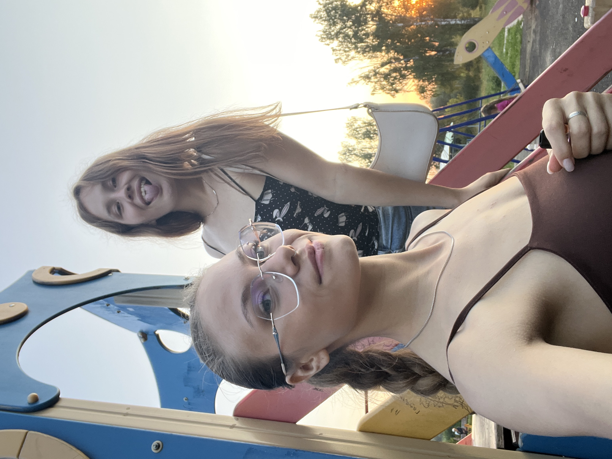 Two young women are posing for a selfie at a playground in the city of Foky, Russia, during sunset. The woman in the foreground is wearing glasses and a brown tank top, and her hair is in a braid. The woman behind her is wearing a black tank top with a bunny print and jeans, and she is sticking her tongue out. In the background, the sun is setting through the trees, casting a warm glow on the scene. A playground structure, consisting of blue and yellow metal components and a red slide, is visible.