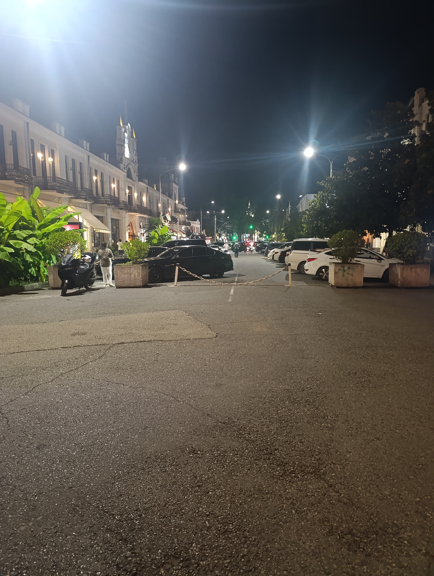 The image depicts a street scene at night in Sokhumi, Abkhazia. The street is paved and appears to be wet, reflecting the artificial lights. On the left side of the street, a building with a white facade and multiple balconies is visible, illuminated by outdoor lights. In front of this building, large potted plants and some awnings are present. A black motorcycle is parked on the side of the street, and a person wearing light-colored pants and a patterned shirt stands nearby. Several black cars are parked along the curb. Further down the street, a line of white vehicles, including a van and sedans, are parked. Trees are visible on the right side of the street, and streetlights cast illumination. The sky is dark, indicating nighttime.