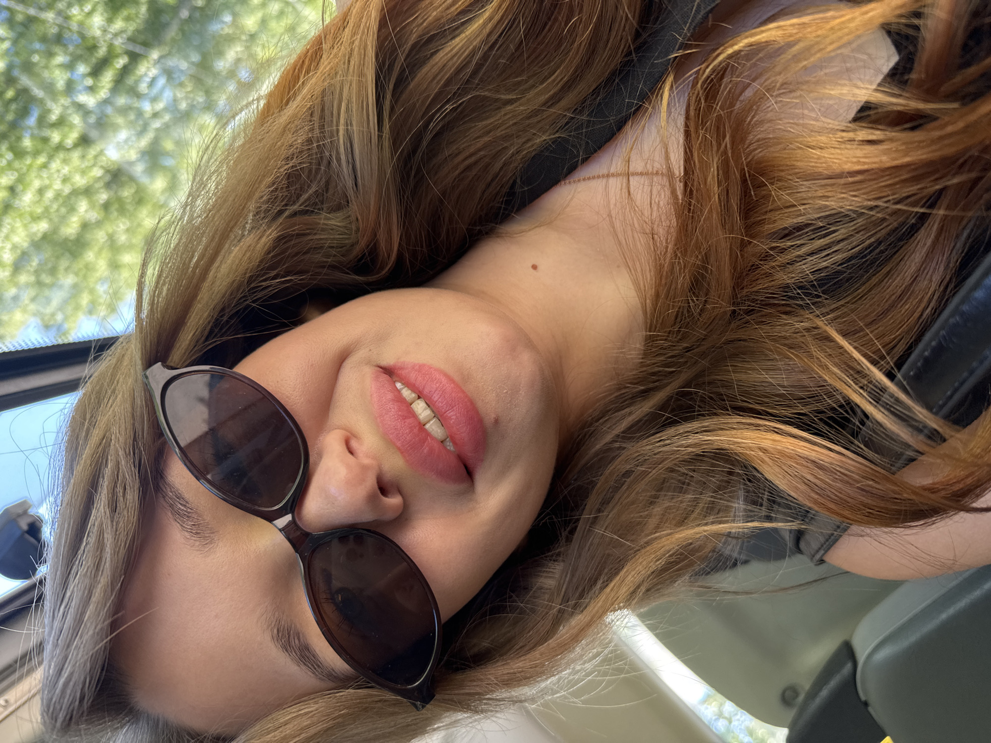 A young woman with long, wavy auburn hair is taking a selfie in what appears to be the interior of a vehicle, likely a car. She is wearing dark, angular sunglasses that obscure her eyes, and her lips are painted a vibrant pink. Her head is tilted slightly, and she is smiling, revealing a hint of her teeth. The background is blurred, but the greenery suggests an outdoor setting with trees and sunlight filtering through. The image is taken from a high angle, looking down at the woman. The scene is consistent with a candid shot taken during a car ride, possibly on a sunny day. There is no specific indication of the city of Chaikovsky, Russia.