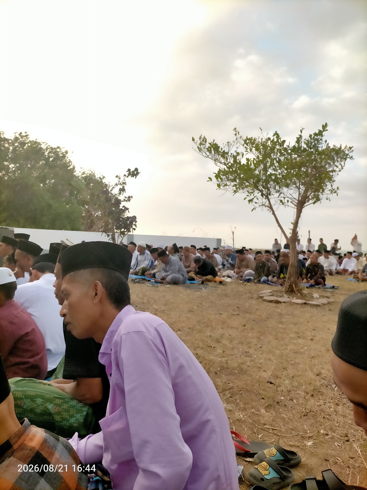 The image depicts a gathering of people, likely men, dressed in traditional Islamic attire, including peci (caps) and shirts, seated on the ground in an outdoor area. They appear to be engaged in a prayer or religious service. The background shows a few trees and a cloudy sky. The ground is covered with dry grass and dirt. In the foreground, the faces of several individuals are visible, some looking towards the distance, others with their heads bowed. The overall setting suggests a communal religious event, possibly Eid al-Fitr or Eid al-Adha, taking place in a rural or semi-urban location in Kranji, Indonesia. A timestamp "2026/08/21 16:44" is visible in the lower-left corner of the image.