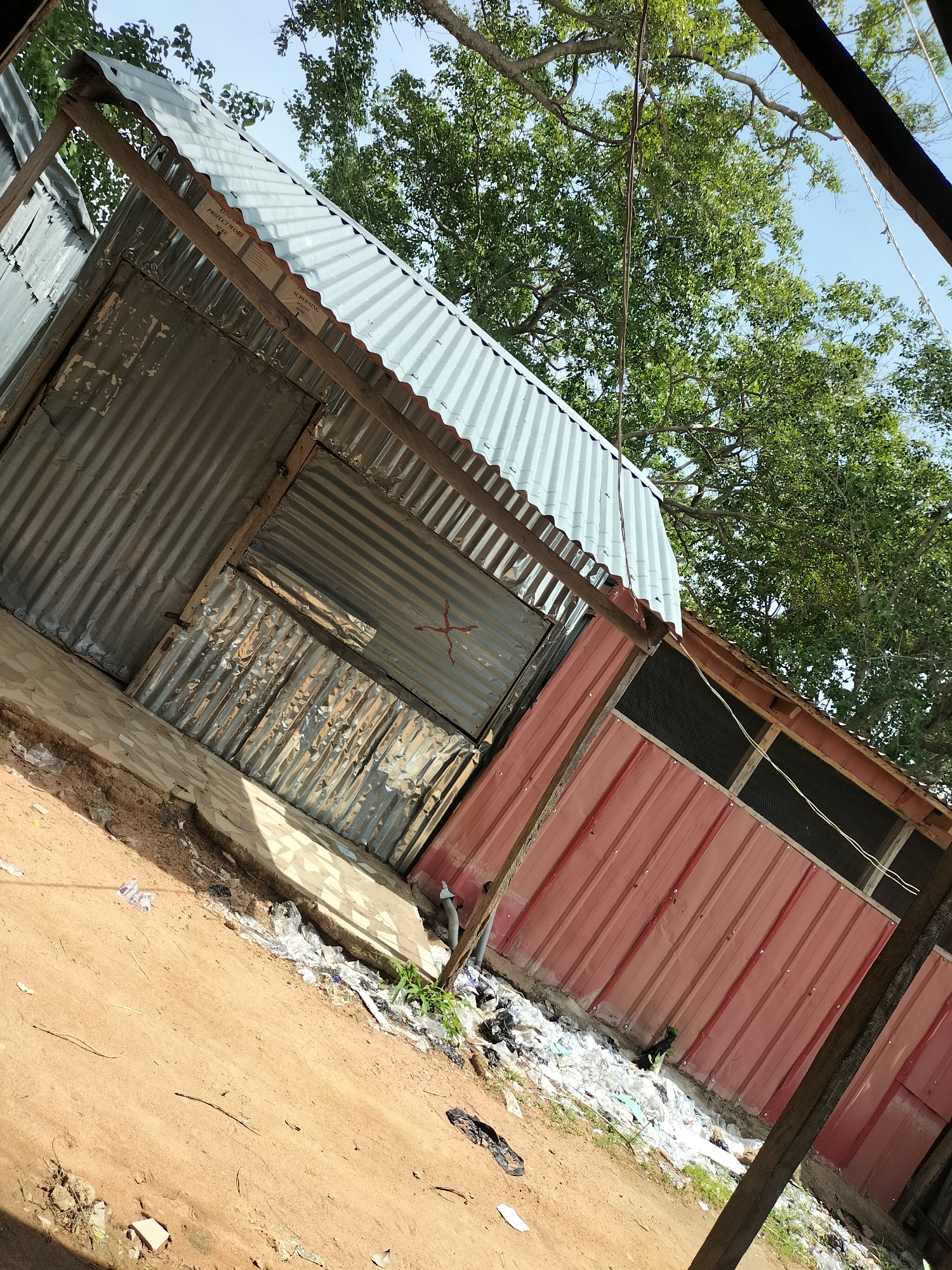 The image depicts an outdoor scene, likely in Jalingo, Nigeria, showing a makeshift structure with a corrugated metal roof and walls. The structure appears to be a stall or a small building with multiple sections. One section has red metal siding, while another is constructed with corrugated aluminum. There are visible wooden supports holding up the roof. The ground in front of the structure is sandy and littered with various types of trash, including plastic bags and other debris. Lush green trees with a clear blue sky are visible in the background, suggesting an outdoor, possibly rural or semi-urban, setting. There are no people visible in the image. The overall impression is one of a functional but unkempt outdoor commercial or storage space.