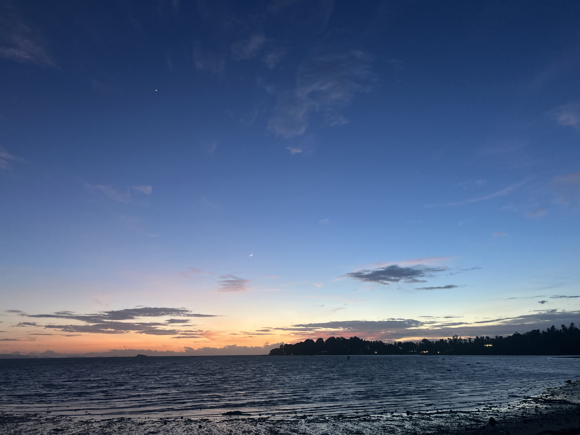 The image depicts a serene twilight scene over a body of water, likely the ocean, near Ko Pha-ngan District, Thailand. The sky transitions from a deep blue at the top to hues of orange and pink near the horizon, indicating either sunrise or sunset. Wispy clouds are scattered across the sky, some catching the fading light. A crescent moon is visible in the upper left quadrant.

In the middle ground, a silhouetted tree line with scattered lights suggests a coastline with some development. The water in the foreground is relatively calm, with visible ripples and texture, and a foreshore or beach area composed of sand, pebbles, and possibly some seaweed. The overall atmosphere is peaceful and tranquil. There are no identifiable people or specific actions taking place.