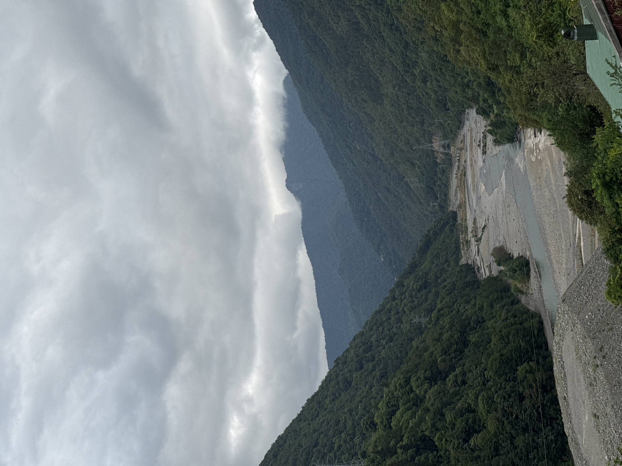 The image shows a mountainous landscape with a river flowing through a valley. The slopes of the mountains are covered with dense green forests. The sky is overcast with dark clouds, suggesting a cloudy or potentially rainy day. In the foreground, a portion of a green roof and chimney is visible, indicating the presence of a building or structure. The riverbed appears to be wide and filled with gravel and stones. There are no people or animals visible in the scene. The overall impression is of a remote and natural environment. The location is identified as Tibeta, Georgia.