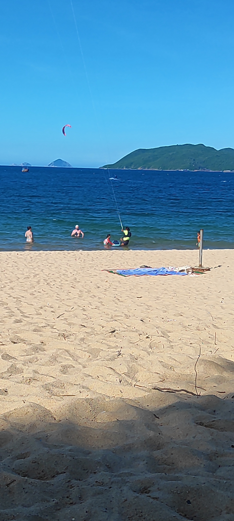 A wide-angle, eye-level shot captures a bright, sunny day at a beach. The foreground is dominated by soft, light brown sand, with some scattered debris. In the middle ground, the turquoise ocean meets the shore, with several people wading in the water. One person, wearing a life vest, appears to be engaged in a watersport, holding onto a kite that stretches up into the clear blue sky. In the distance, a few boats are visible on the water, and a green, tree-covered mountain range forms the horizon under the expansive sky. A small island is visible to the left of the main mountain range. On the sand, near the water's edge, a blue towel and some other items are laid out.